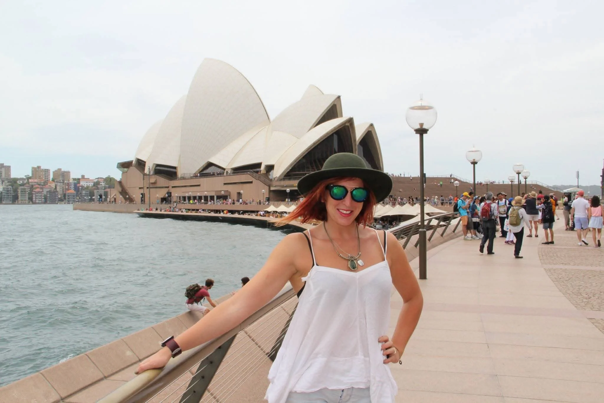 A woman in a white summer dress and hat standing near the edge of a waterfront promenade with the Sydney Opera House and Harbour Bridge in the background during daytime.