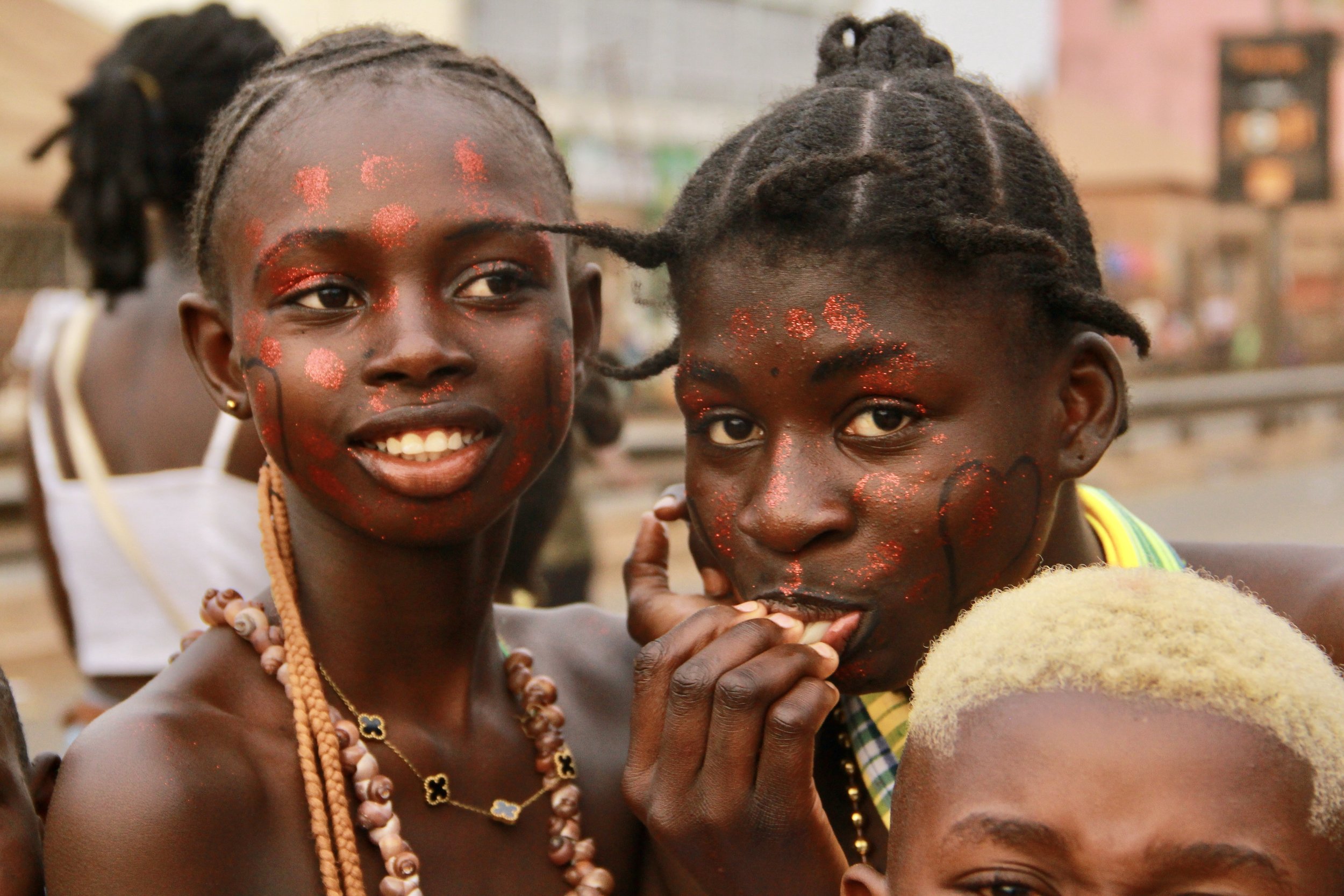 Two young women with face paint and jewelry in an outdoor setting, one smiling and the other with finger near mouth, with a blurred background of people and buildings.