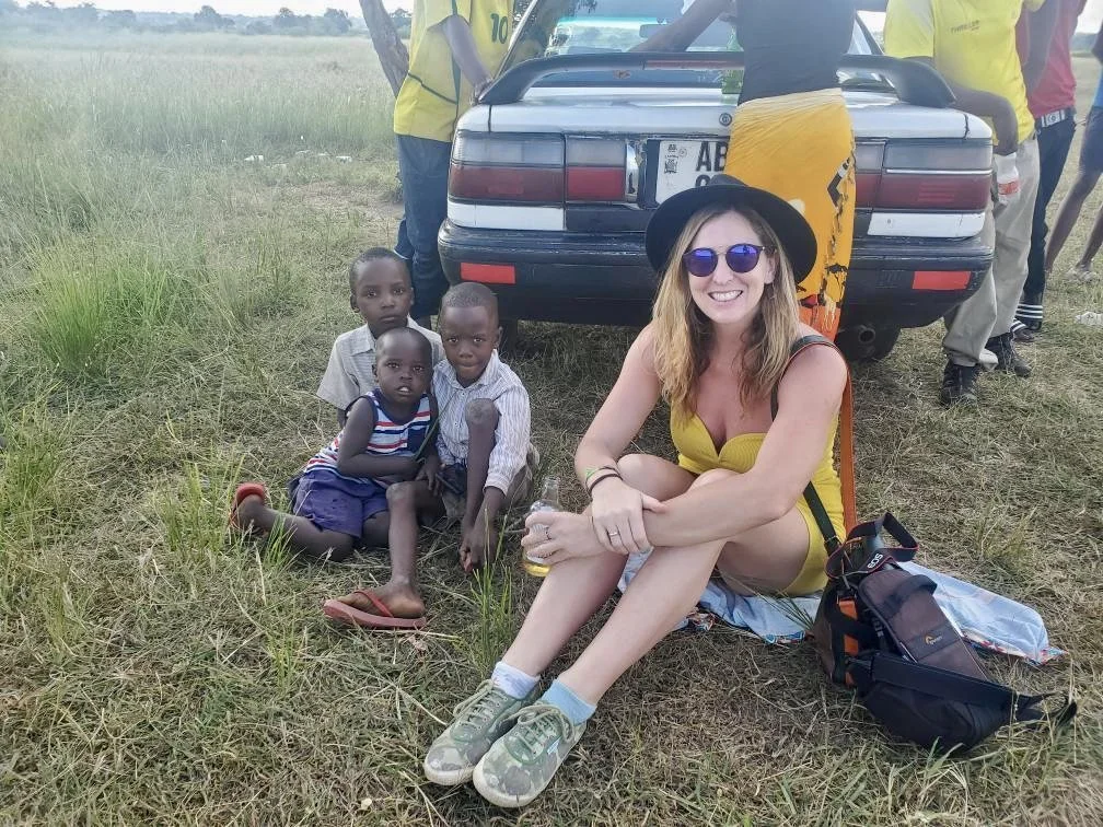 A woman with sunglasses and a black hat sitting on a blanket on the grass, surrounded by four children, with a car parked behind them and several people standing nearby in a grassy outdoor area.