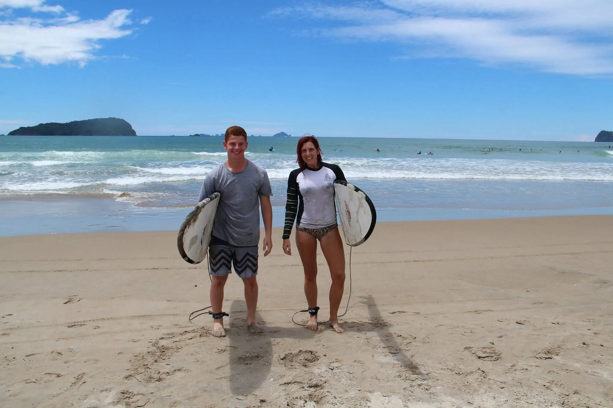A young man and woman standing on a sandy beach, each holding a surfboard, smiling with ocean waves and distant islands in the background.