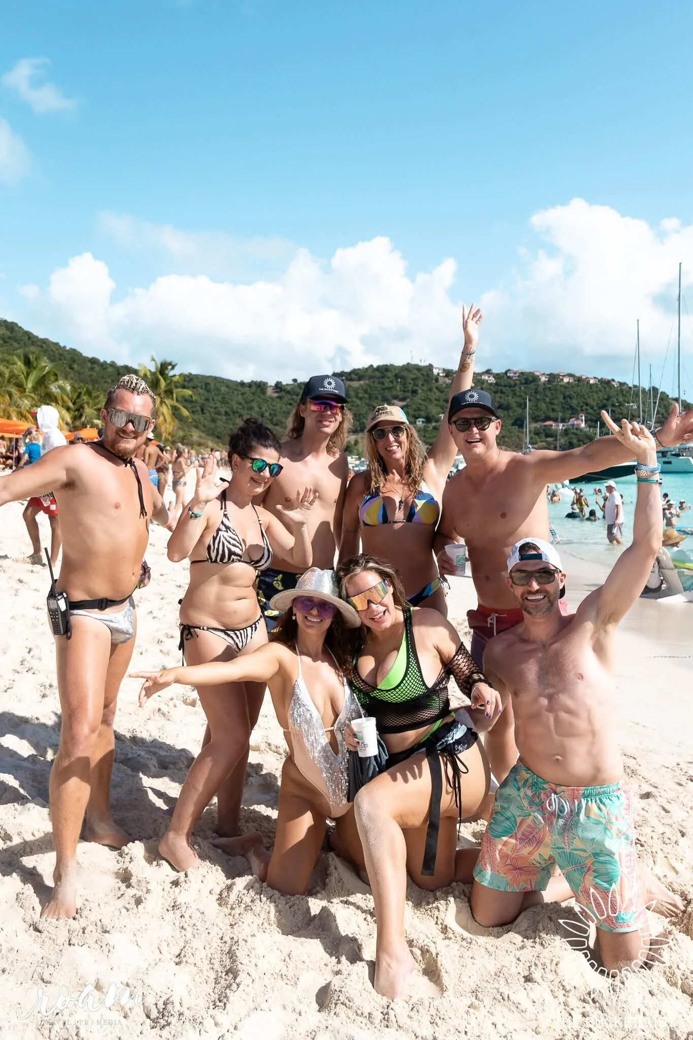 Group of people enjoying a sunny day at the beach, smiling, wearing swimsuits and sunglasses, with boats in the background and a lush green hillside.