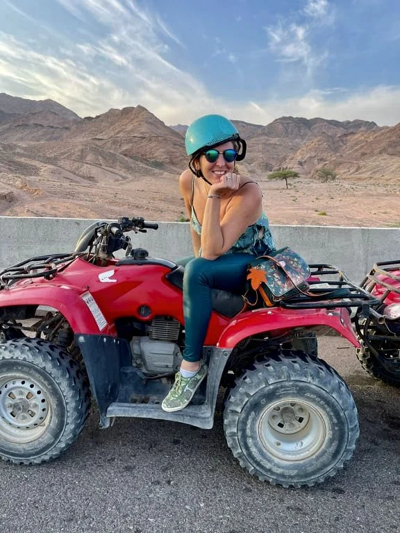 A woman wearing a blue helmet, sunglasses, and workout clothes sits on a red all-terrain vehicle (ATV) in a desert landscape with mountains in the background.
