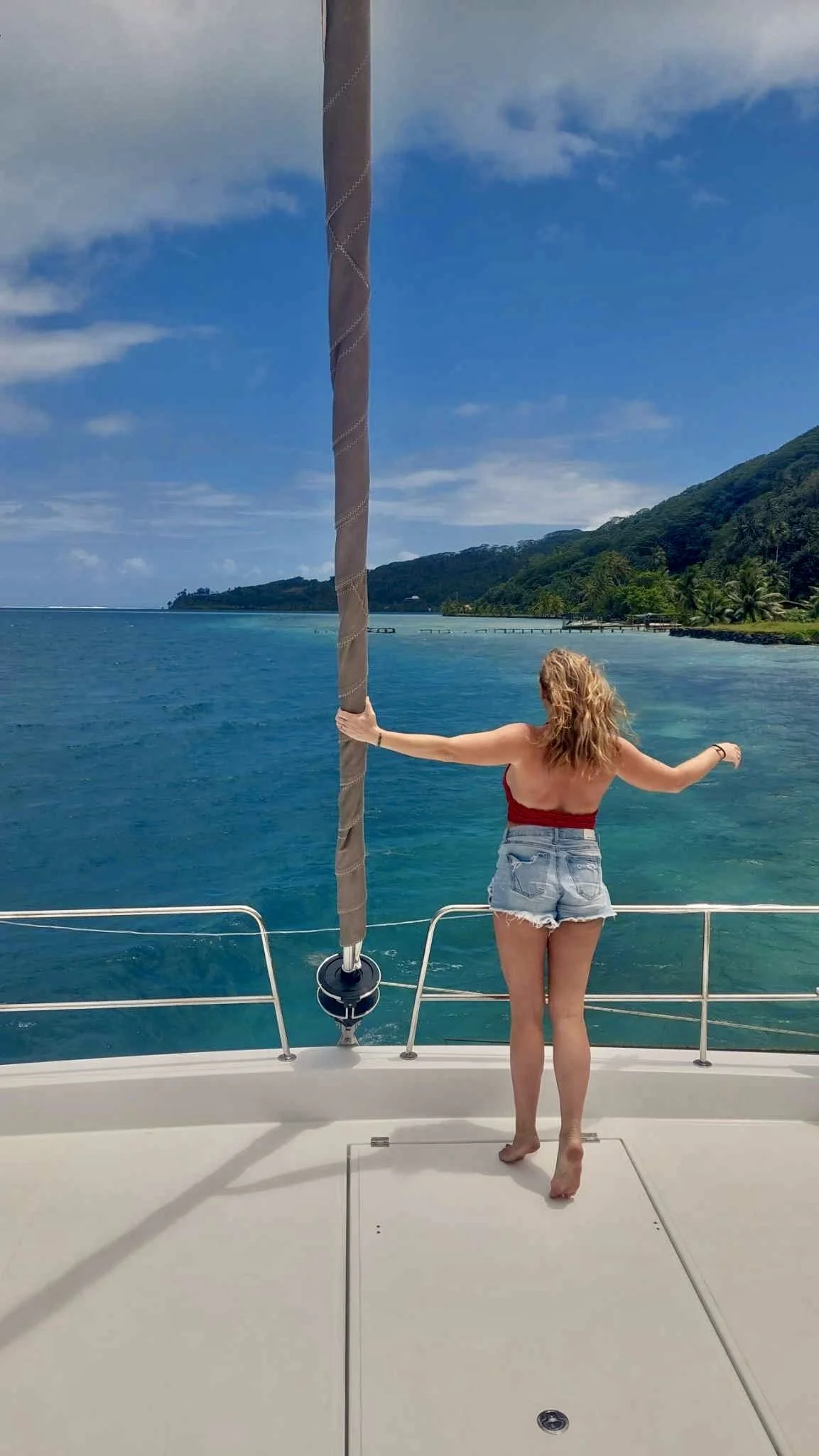A woman with red hair in a red top and denim shorts standing on a boat, holding onto a knot on the sail, looking out at the ocean with a green island in the distance.