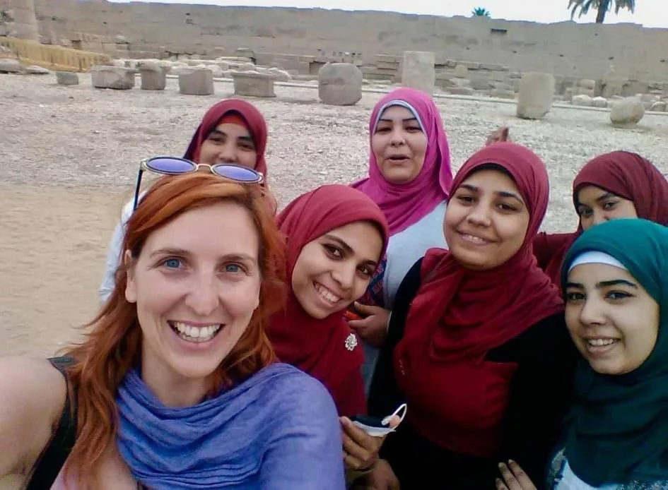 A group of seven women taking a selfie in a desert landscape with stone ruins and palm trees in the background, some wearing colorful headscarves.
