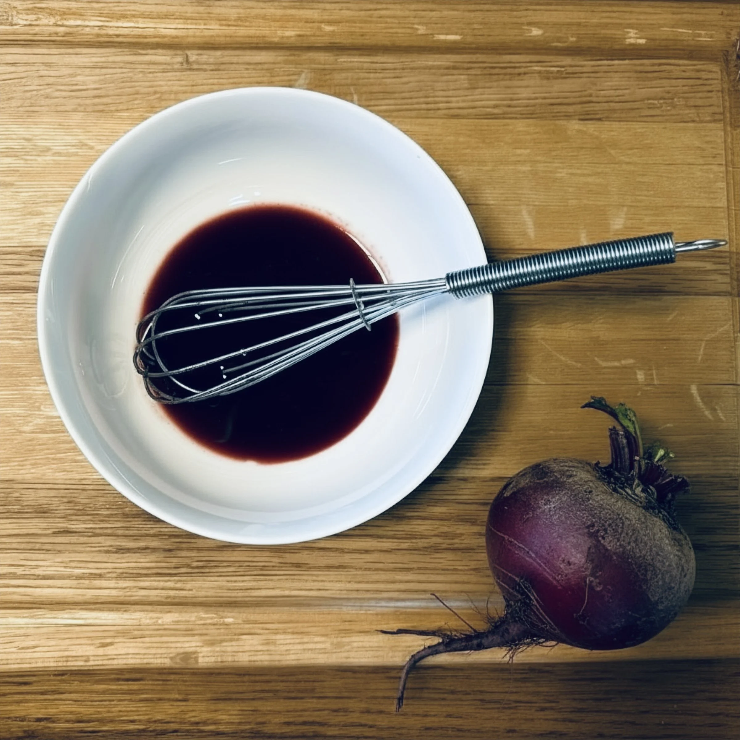 White bowl containing a dark liquid with a whisk inside, placed on a wooden surface. Nearby is a dirt-covered beetroot with green leaves.