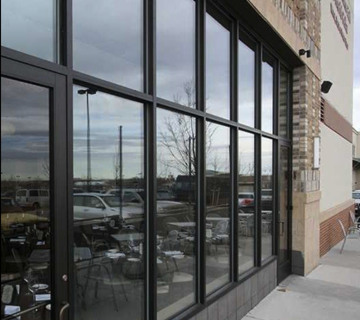 Glass storefront window of a building, reflecting parked cars and cloudy sky outside.