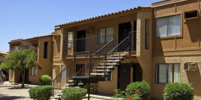 Exterior view of a two-story apartment building with concrete stairs, balcony railings, and several windows. Landscaping includes small bushes and trees.
