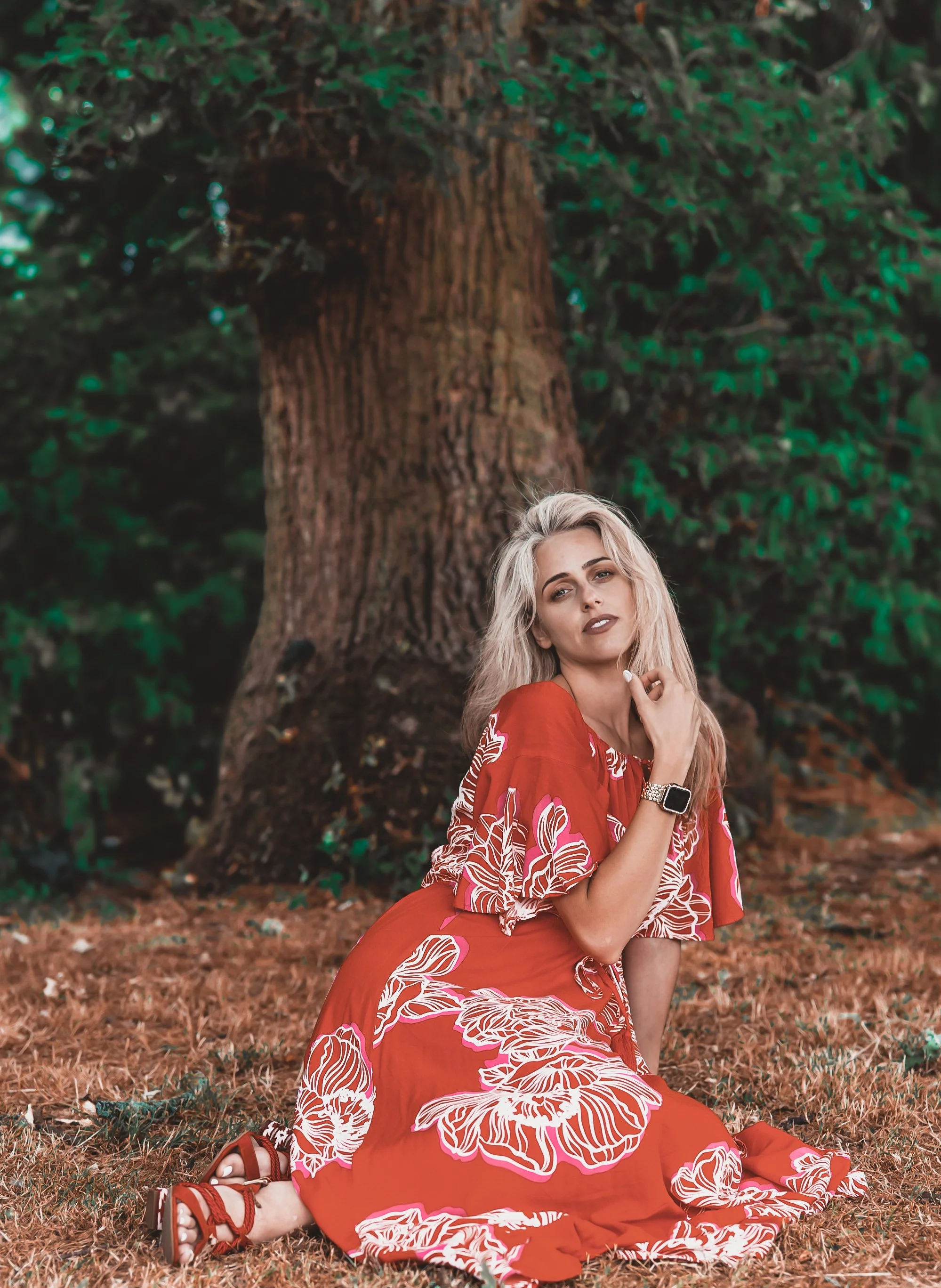 A woman with blonde hair, wearing a red floral dress, sitting on the ground near a large tree in a forested area.