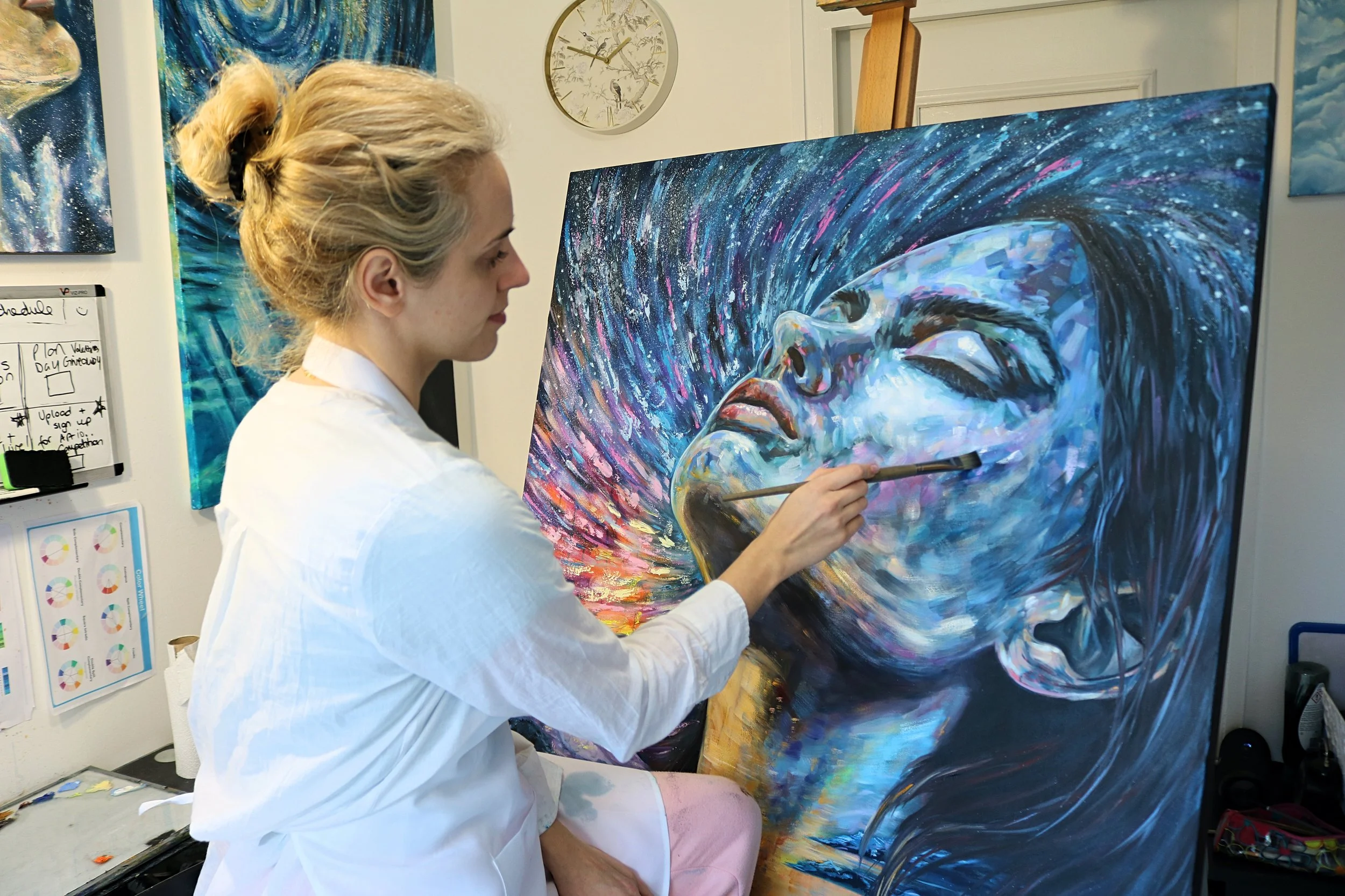 A woman with blonde hair tied back wearing a white shirt painting an abstract portrait of a woman on a large canvas. The painting features vibrant swirls and streaks of blue, white, pink, and black.