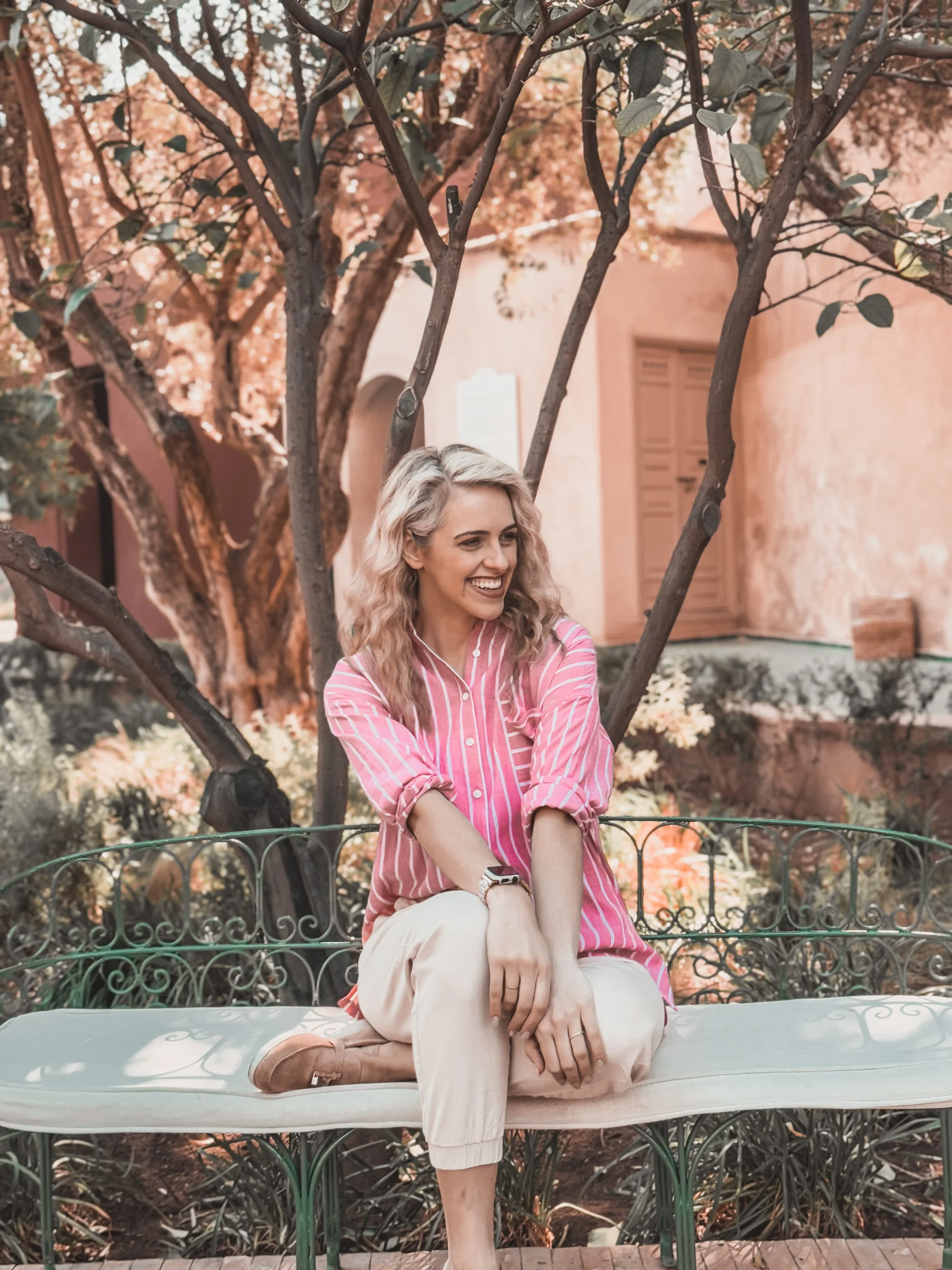 A woman with blonde, curly hair sitting on a white bench outdoors, smiling, wearing a pink and white striped shirt and beige pants, surrounded by trees and pinkish-orange buildings.