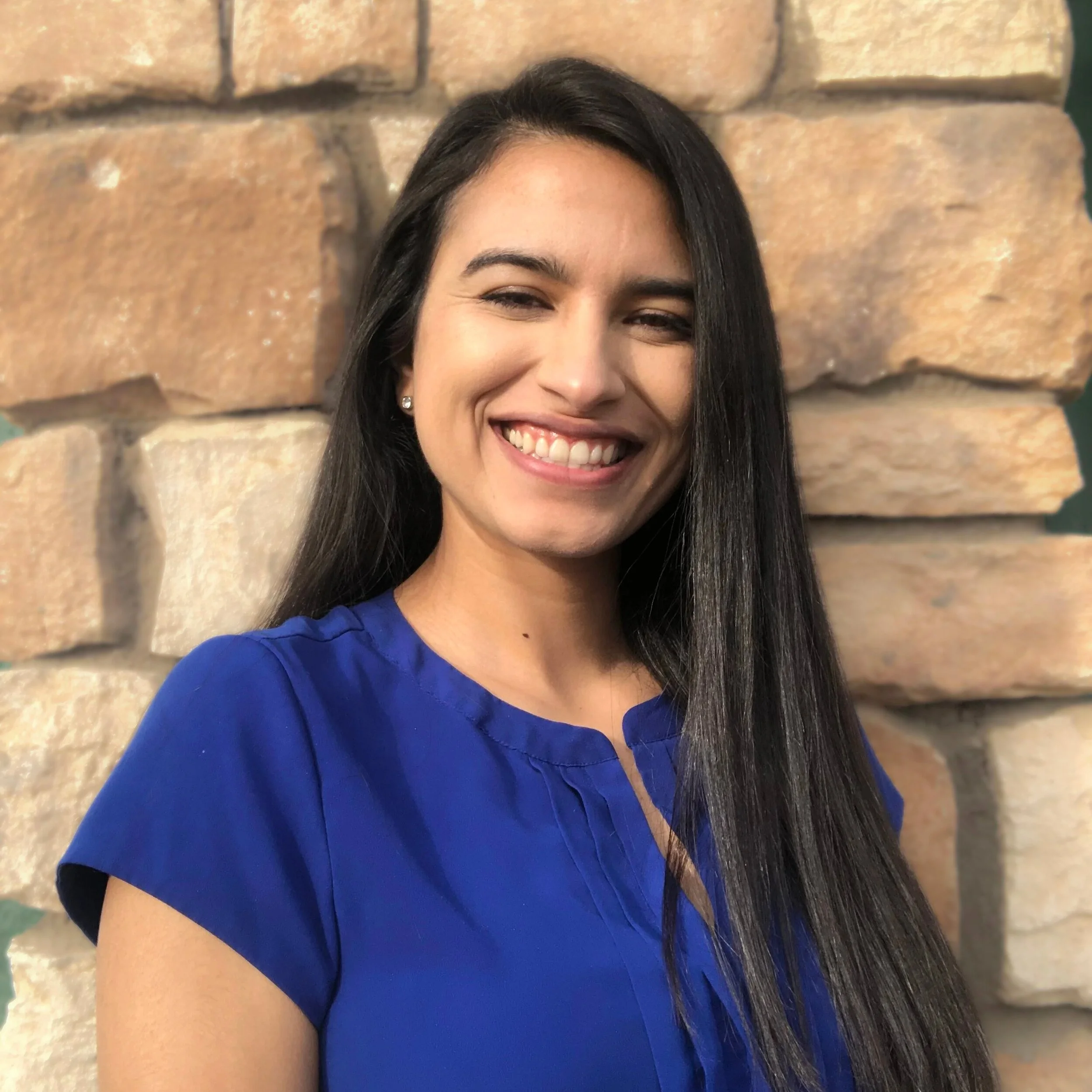 A woman with long black hair smiling, wearing a blue shirt, standing in front of a brick wall.