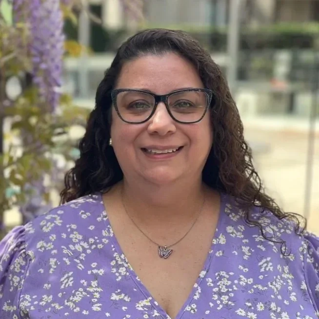 A woman with long curly hair, wearing glasses, a purple floral dress, and a butterfly necklace, smiling outdoors with purple flowers in the background.