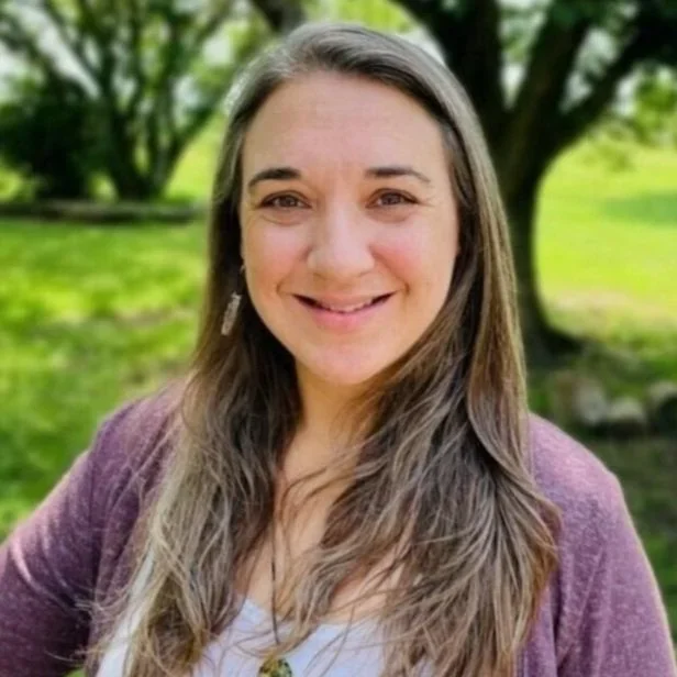 A woman with long brown hair smiling outdoors with green trees in the background.