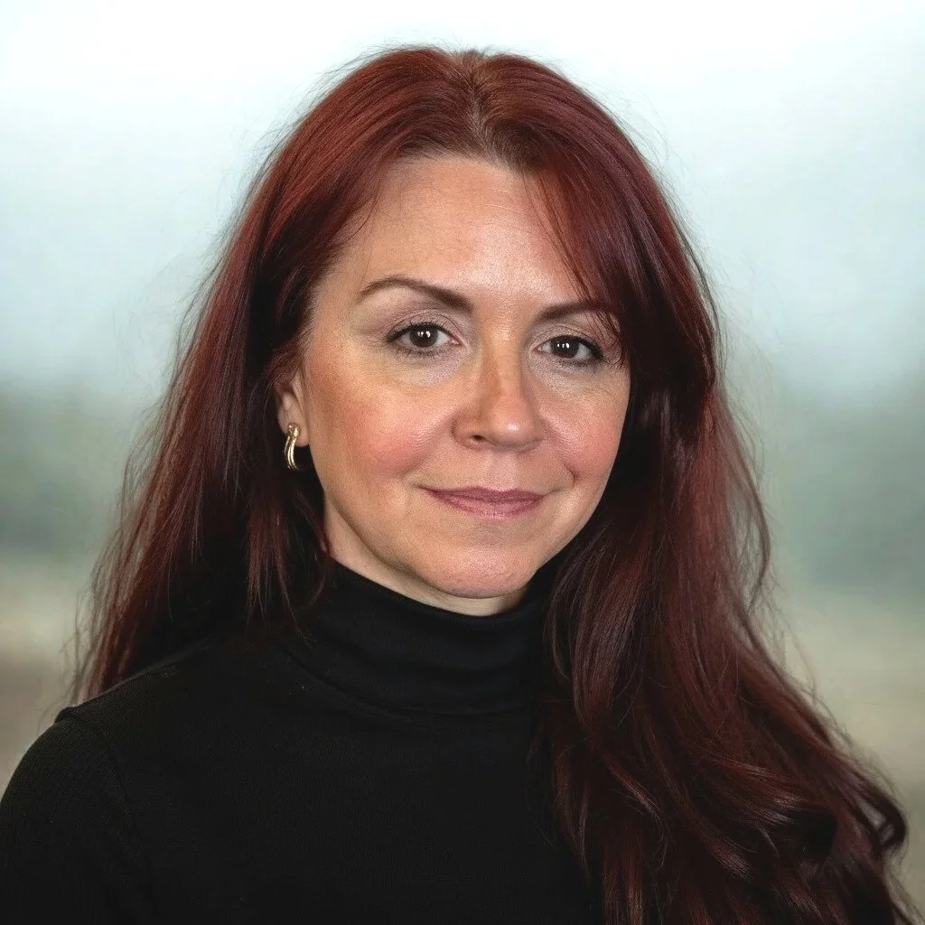 A woman with red hair wearing a black turtleneck and gold hoop earrings, smiling against a blurred outdoor background.
