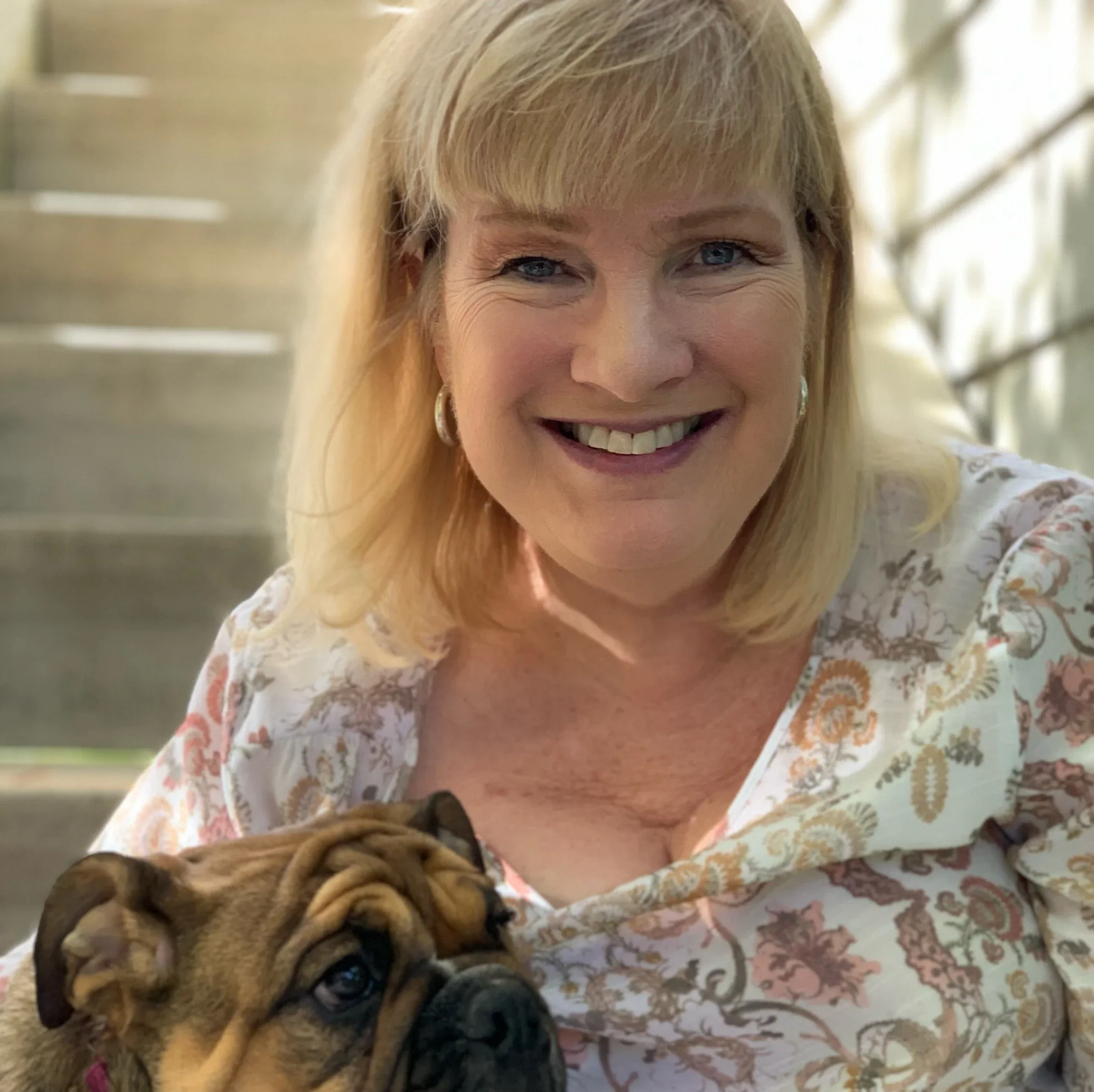 A smiling woman with blonde hair and blue eyes taking a selfie with a small bulldog puppy in her arms. The background is blurred with wooden structures.
