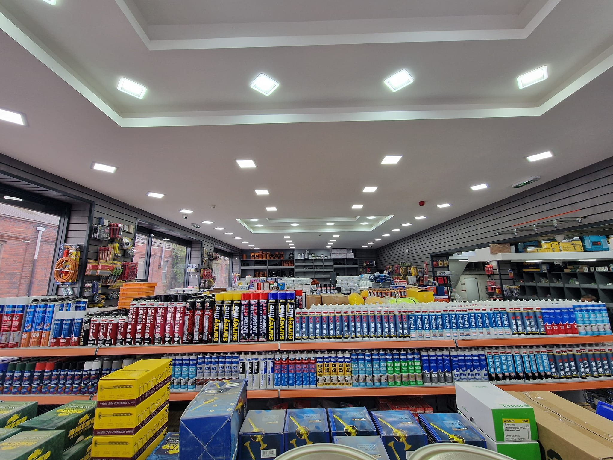 Interior of a hardware or office supply store with shelves stocked with glue, paint, and other supplies, illuminated by ceiling lights.