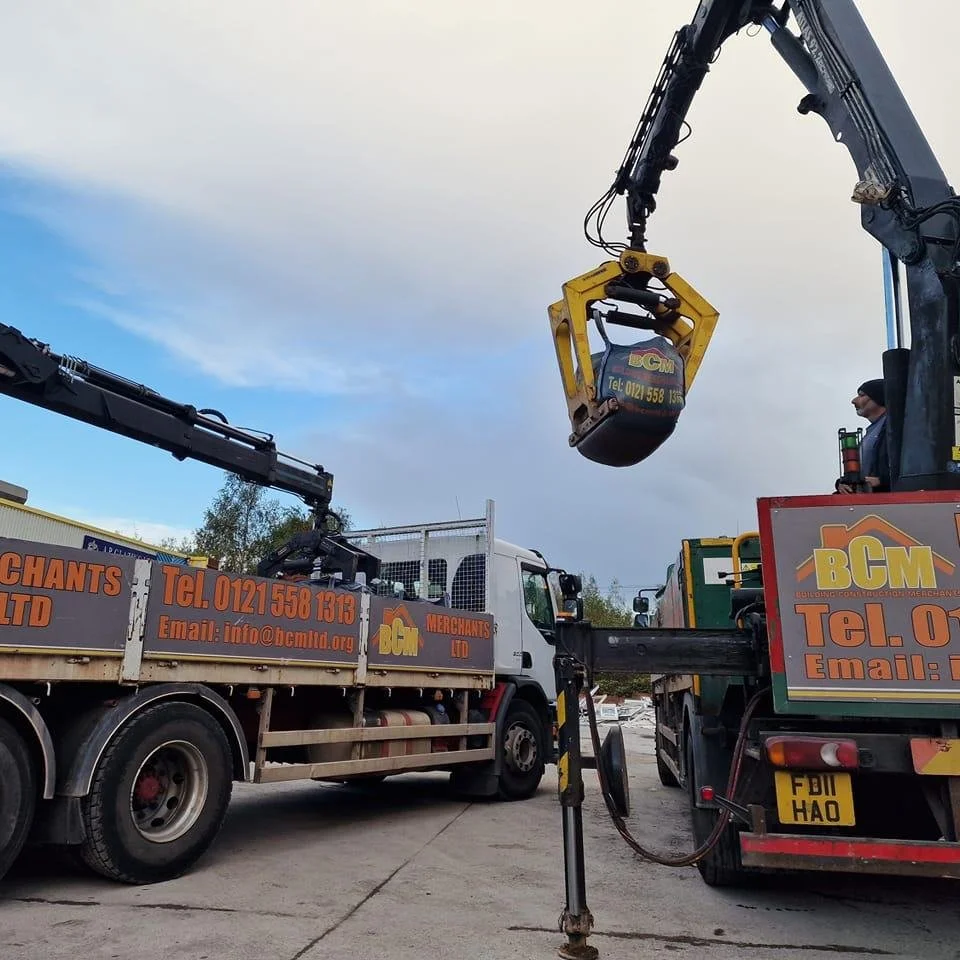 Two trucks are parked with crane equipment and a worker operating the crane. The crane is lifting a small container or device, and the trucks belong to a company called BC M with contact information on the side.