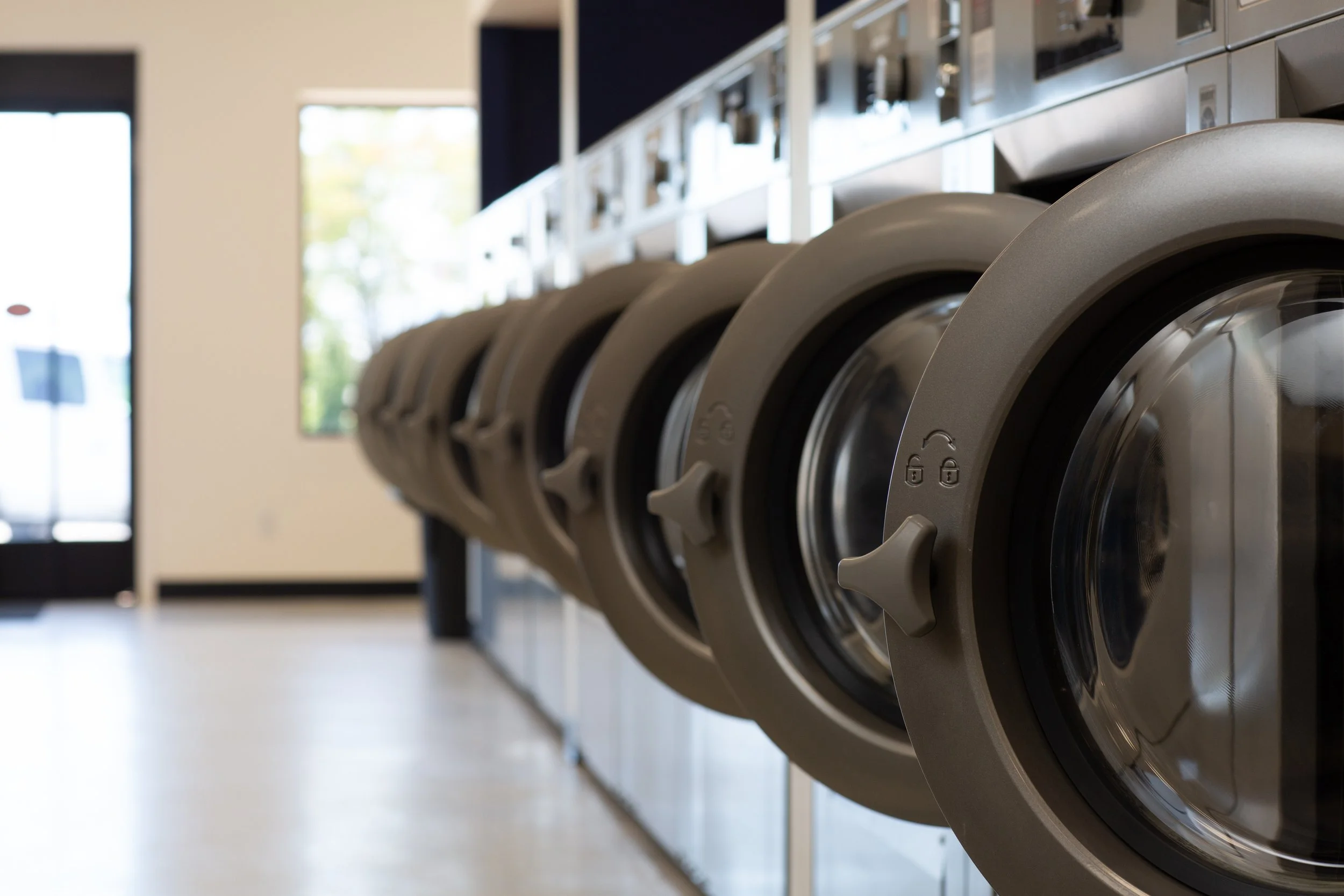 A row of front-loading washing machines in a laundromat, with glass doors and control knobs, lined up against a wall with a window in the background.