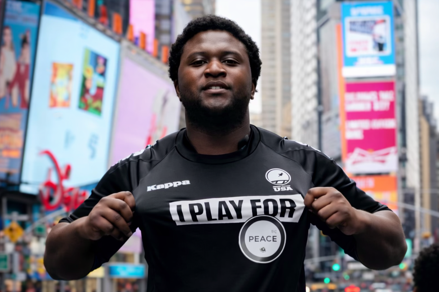 Man in black sports shirt with logos holding up the shirt on a city street with bright digital billboards.
