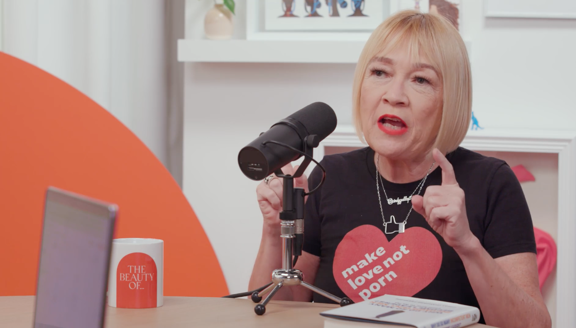 A woman with short blonde hair speaking into a microphone during a podcast or interview. She is wearing a black T-shirt with a red heart and text that says 'make love not porn'. She is gesturing with her right hand, pointing her index finger up, and has a serious expression. There is a cup on the table that says 'THE BEAUTY OF...' and a laptop in front of her.