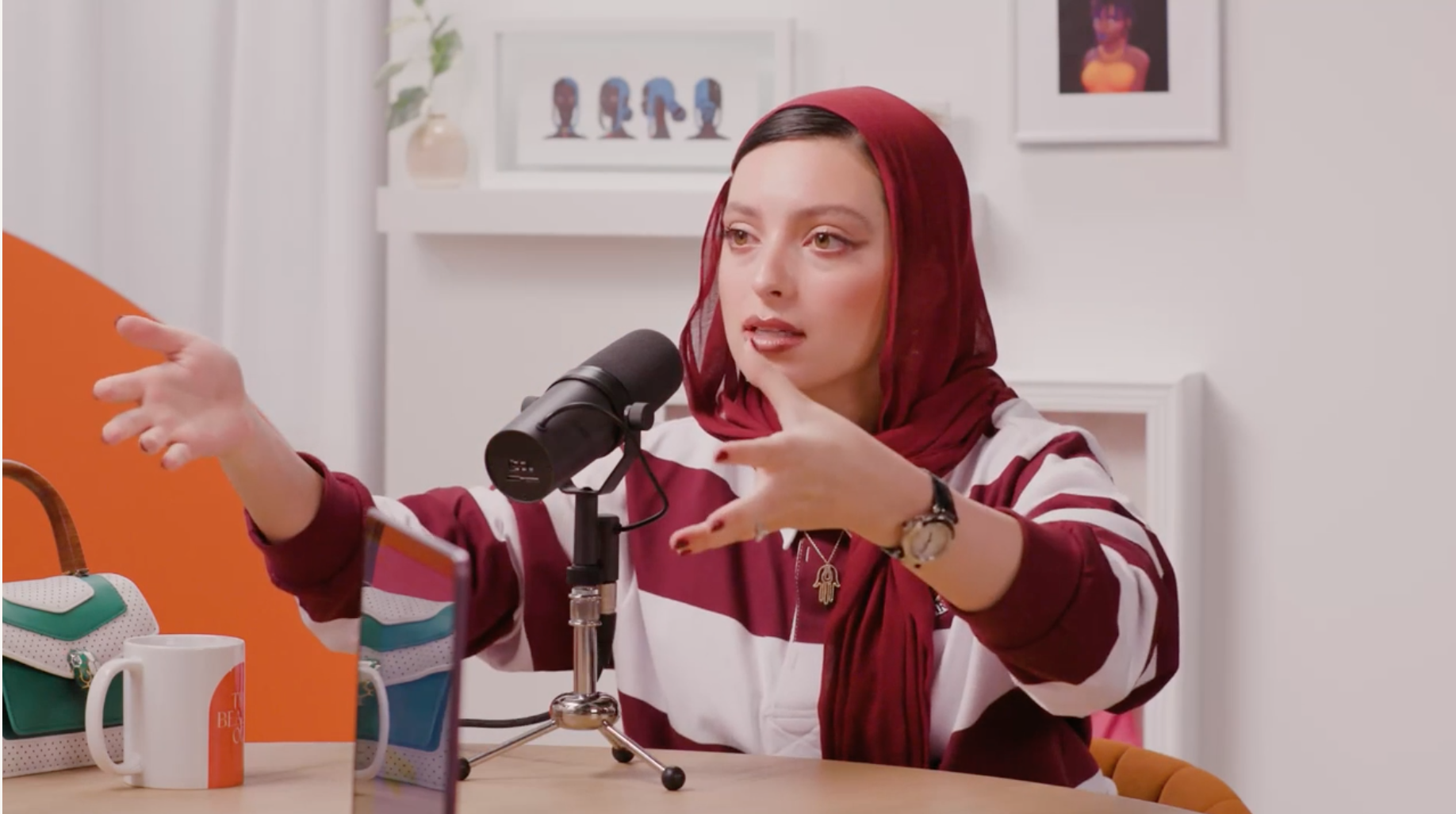 A woman with a red headscarf speaking into a microphone, gesturing with her hands during a podcast or interview in a bright room with artwork and a shelf in the background.