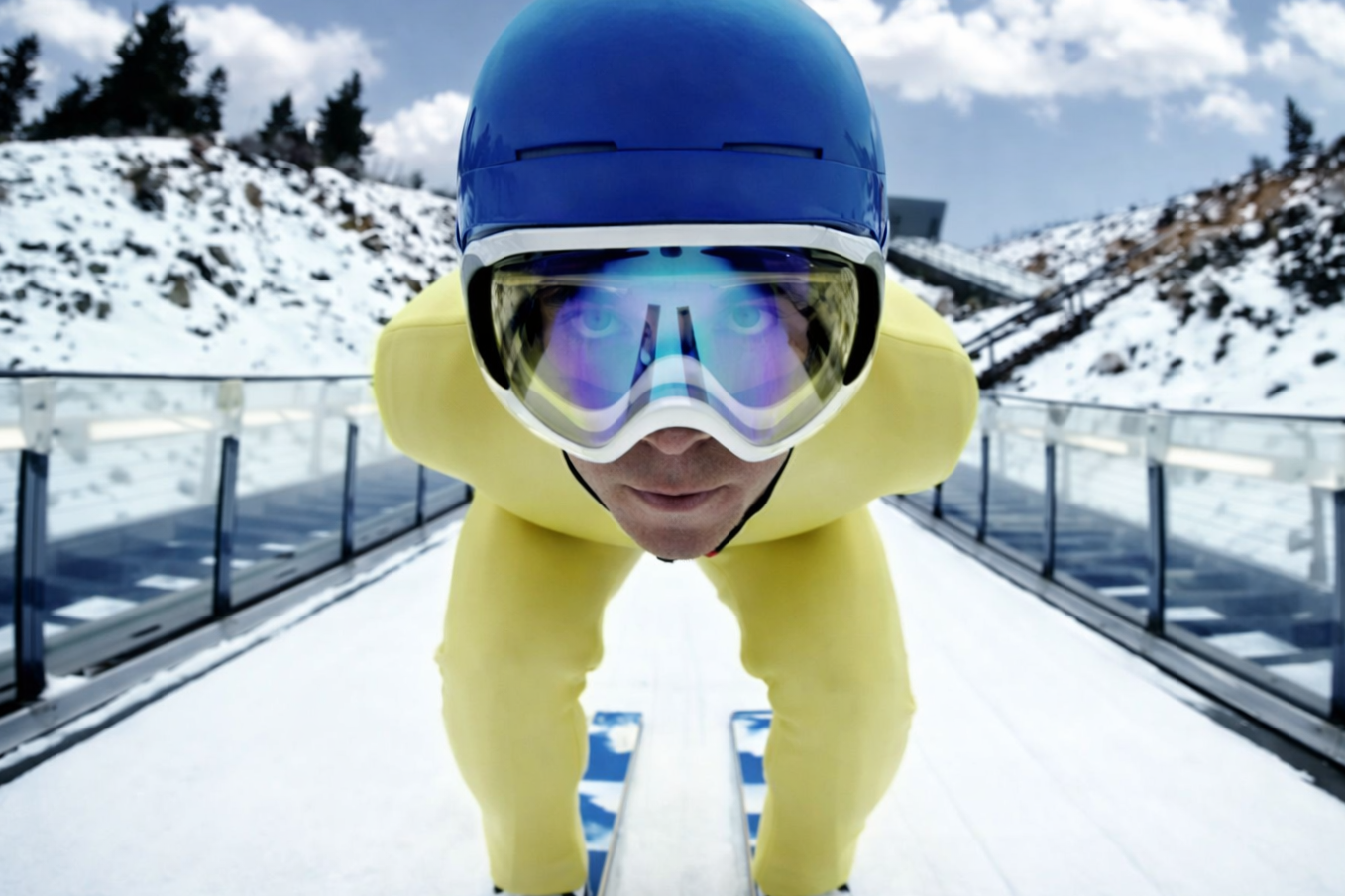 Close-up of a ski jumper in yellow suit and blue helmet, crouched on skis on a snowy slope, with mountains and cloudy sky in background.