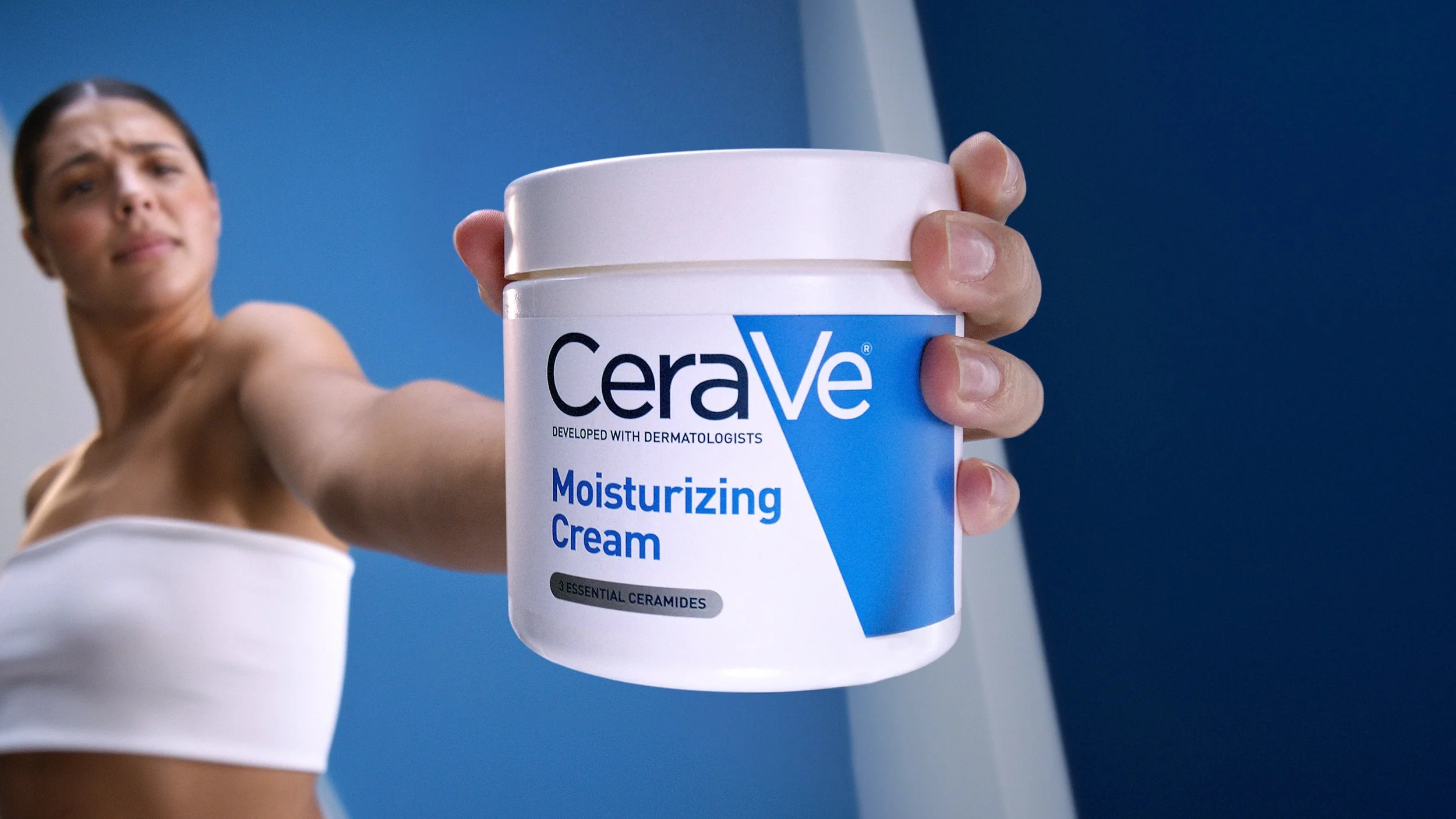 A woman holding a jar of CeraVe moisturizing cream towards the camera. The woman is wearing a white strapless top and is in a room with blue and white walls.