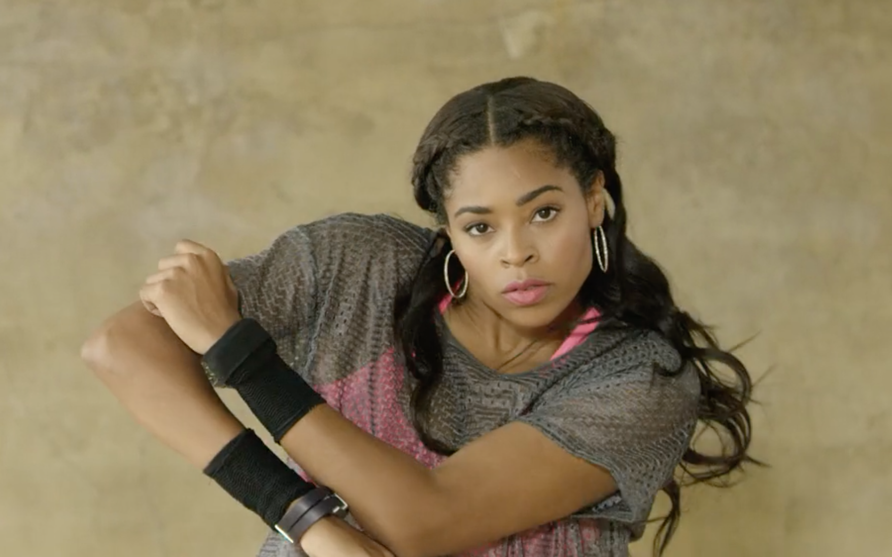 A woman with long dark hair, wearing hoop earrings and a gray and pink sports shirt, holding her right arm with her left arm, looking at the camera with a serious expression.