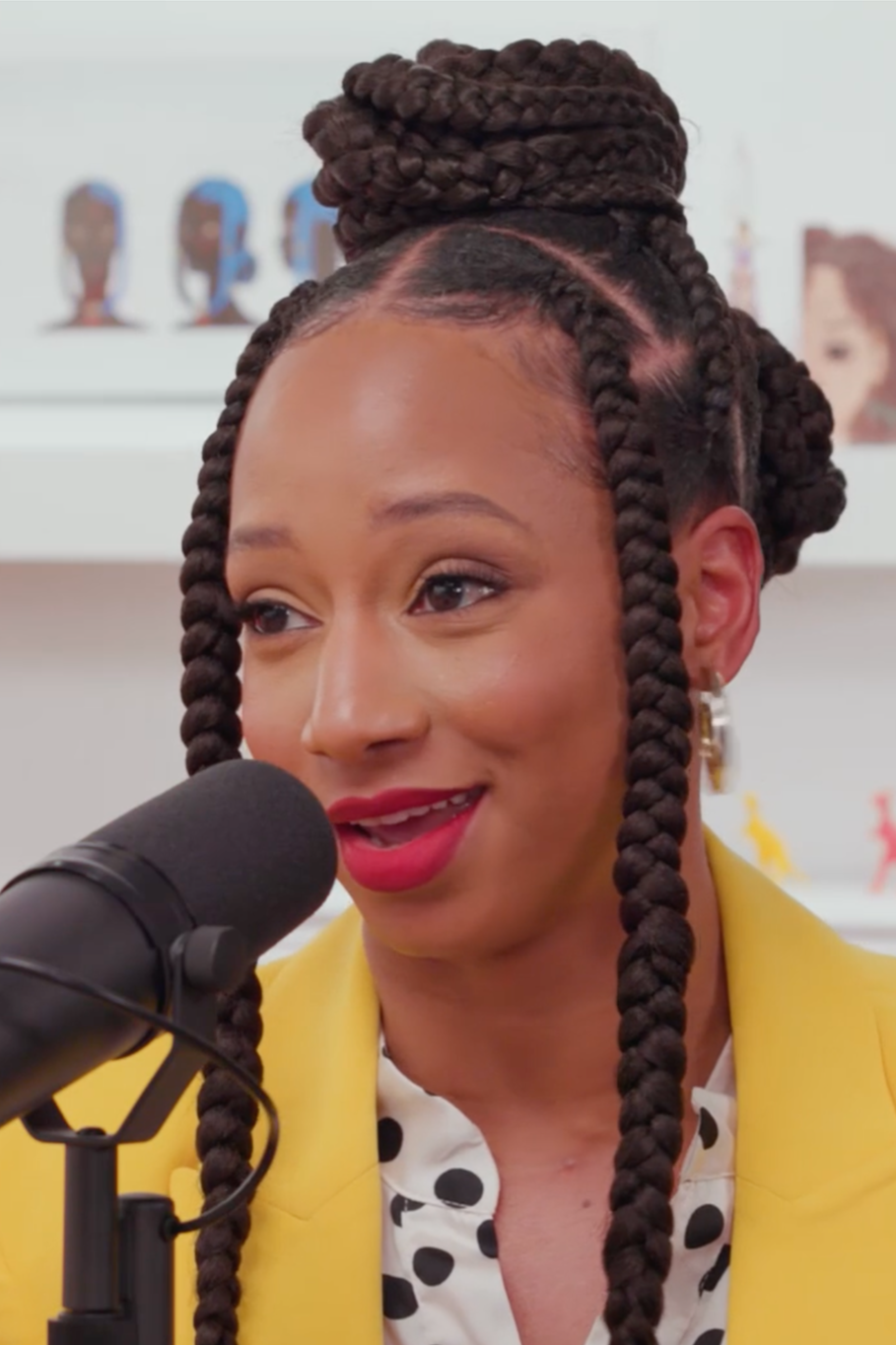 A woman with long twisted braids styled in an updo, speaking into a microphone, wearing a yellow blazer and a white polka-dot blouse, with colorful artwork in the background.