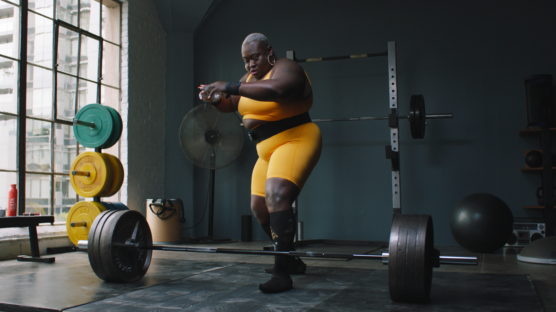 A woman in yellow workout attire is preparing to lift a barbell with weights in a gym. The gym has large windows, weight plates, and workout equipment around.