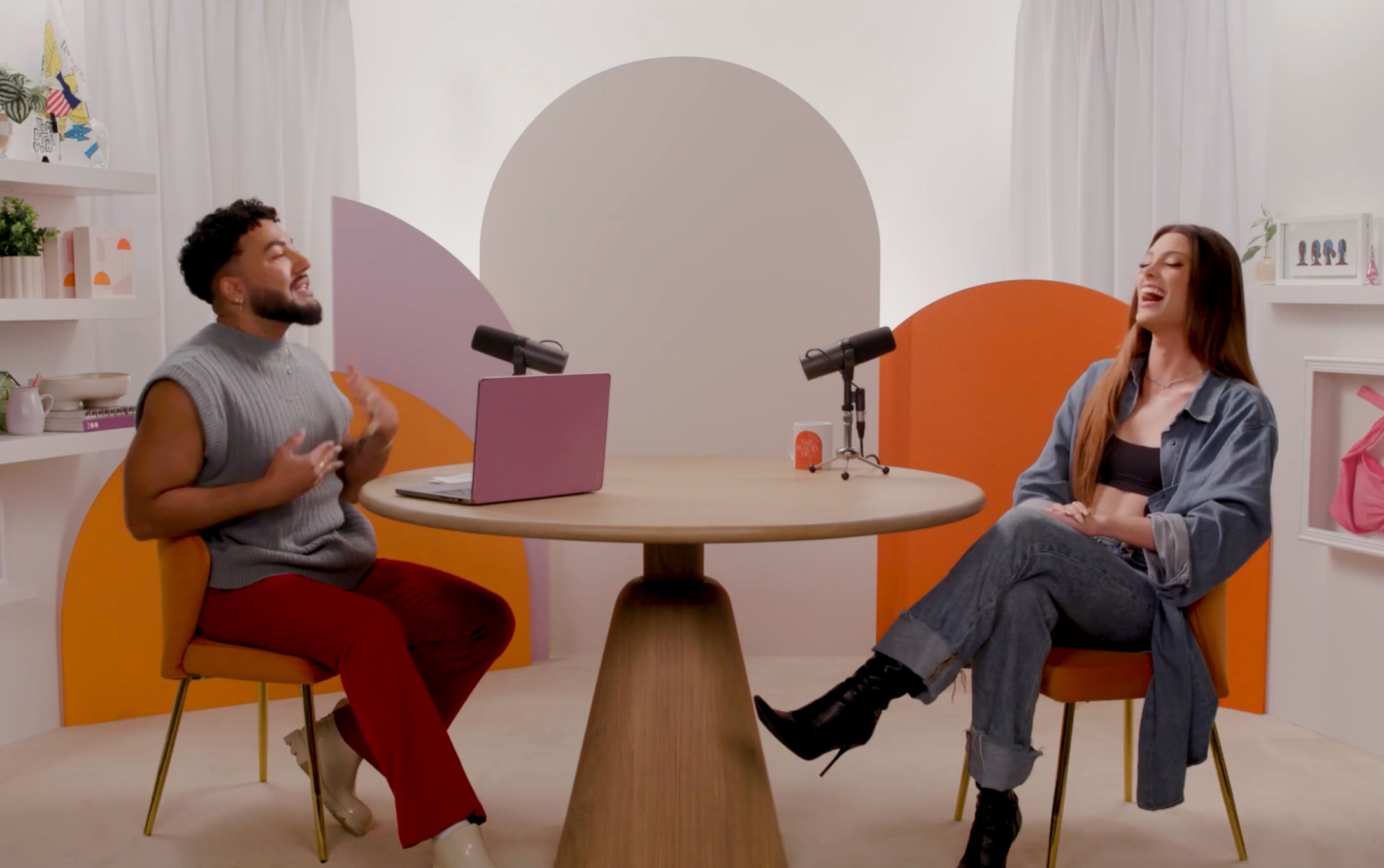 Two people are sitting and talking at a round table with a pink laptop and two microphones, in a colorful room with abstract wall art and shelves.