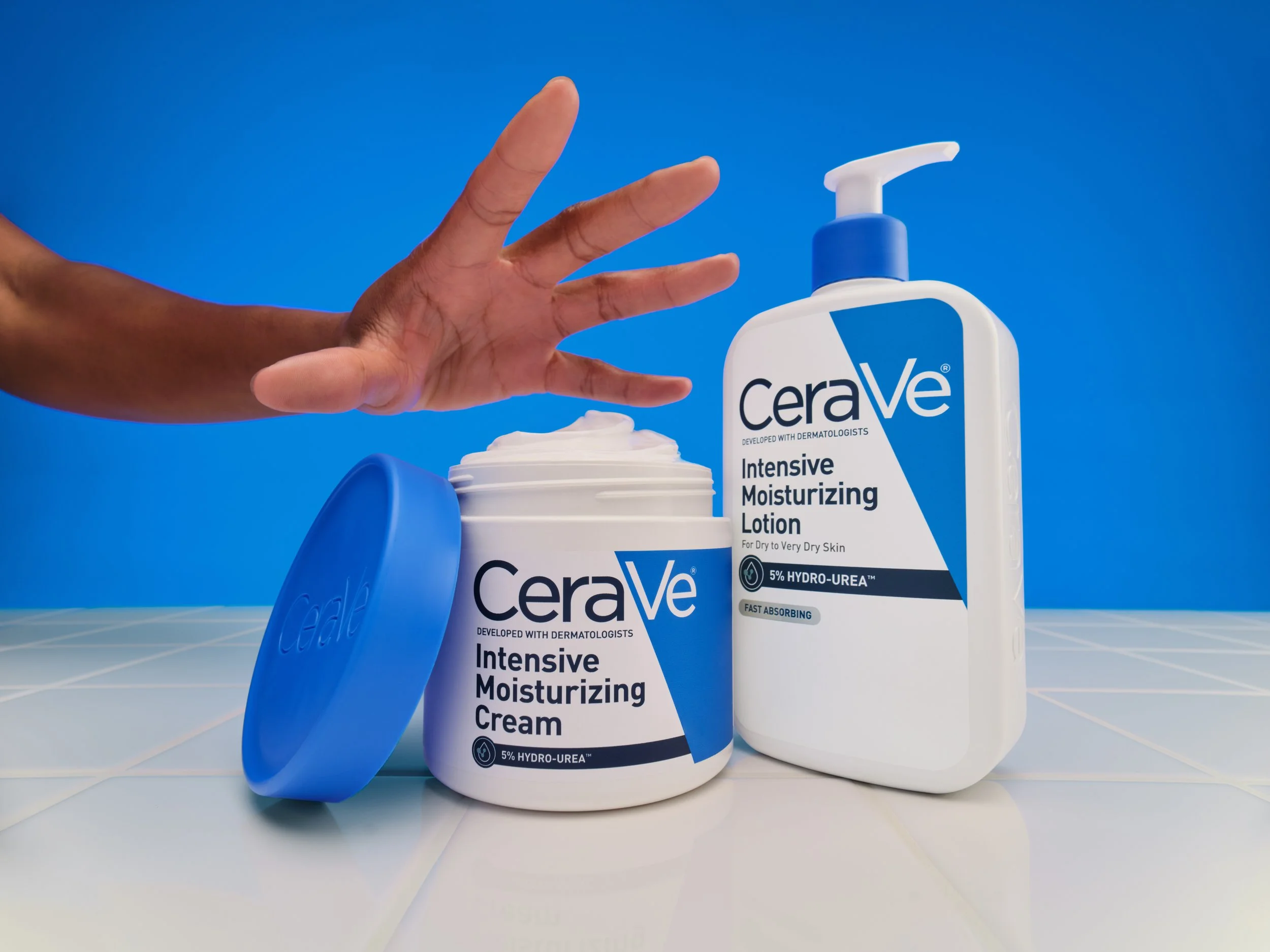 A hand reaching towards skincare products: a jar of CeraVe Intensive Moisturizing Cream with its lid off, and a bottle of CeraVe Intensive Moisturizing Lotion with a pump dispenser on a tiled surface with a blue background.