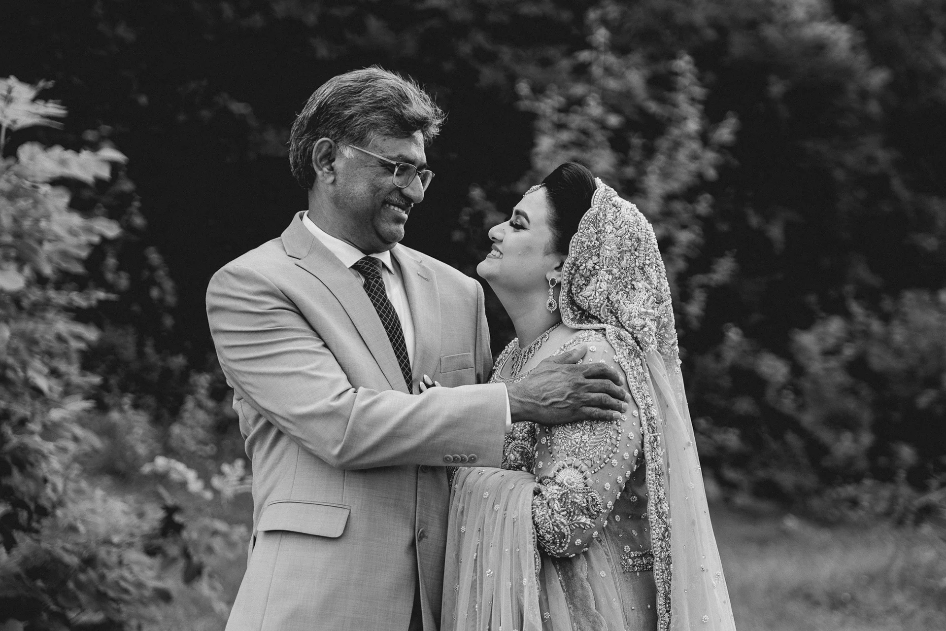 A man and woman are smiling and looking at each other outdoors, the man in a light-colored suit and the woman in an ornate traditional dress and head covering.