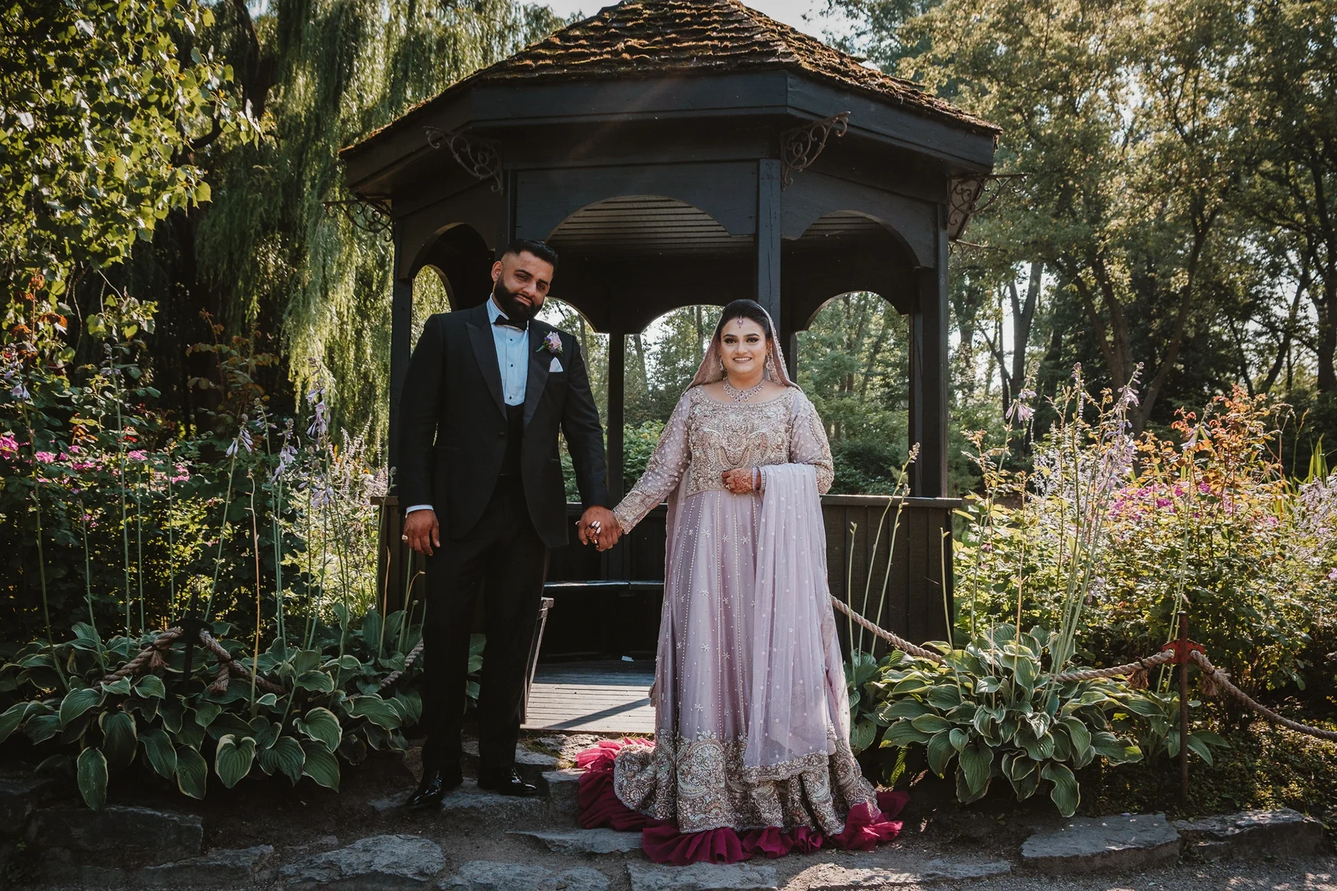 A newlywed couple holding hands and standing in front of a small wooden gazebo in a garden, with trees and colorful flowers surrounding them.