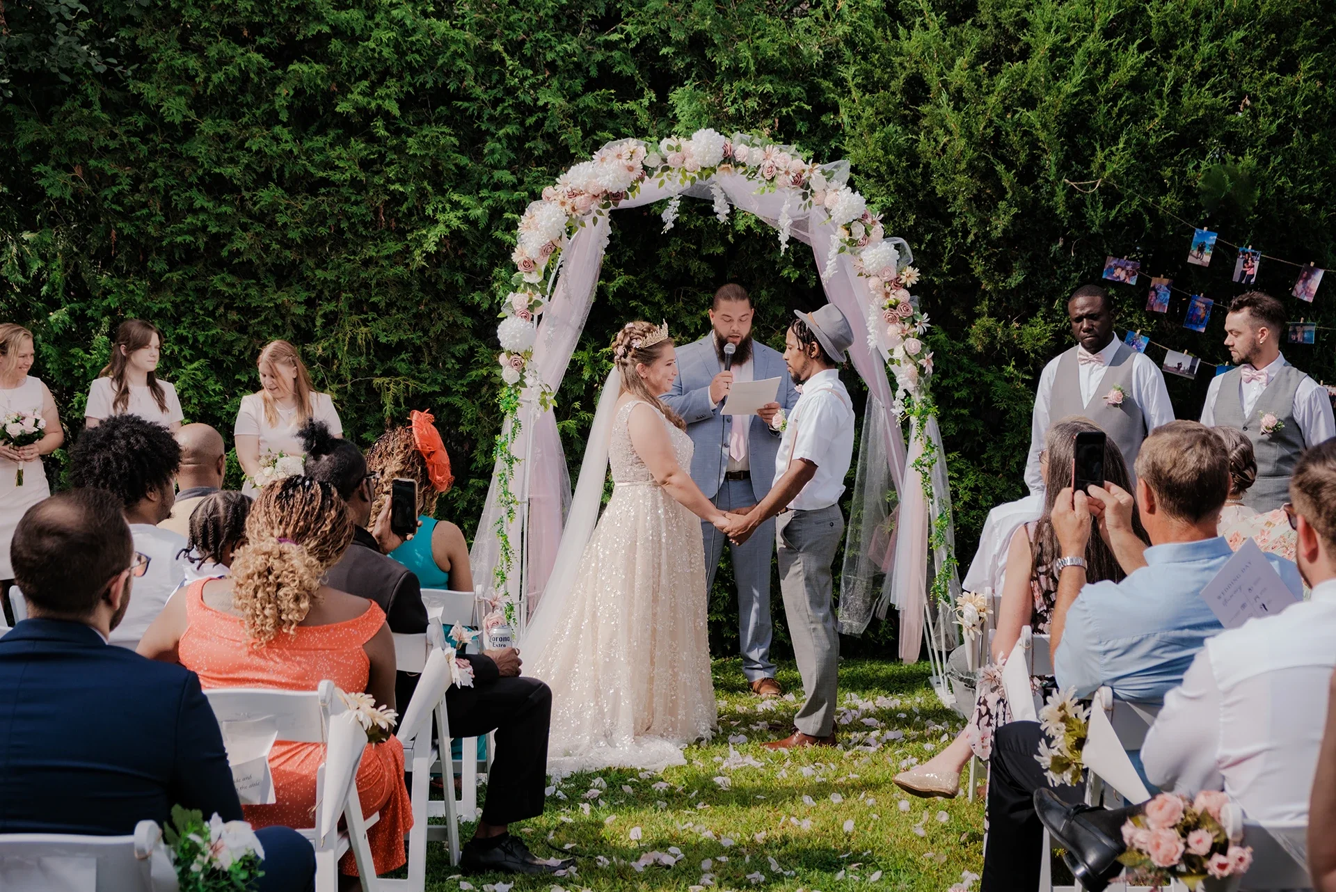 A wedding ceremony outdoors with a bride and groom holding hands under a floral arch, surrounded by guests and bridesmaids on one side, groomsmen on the other, with onlookers taking photos.