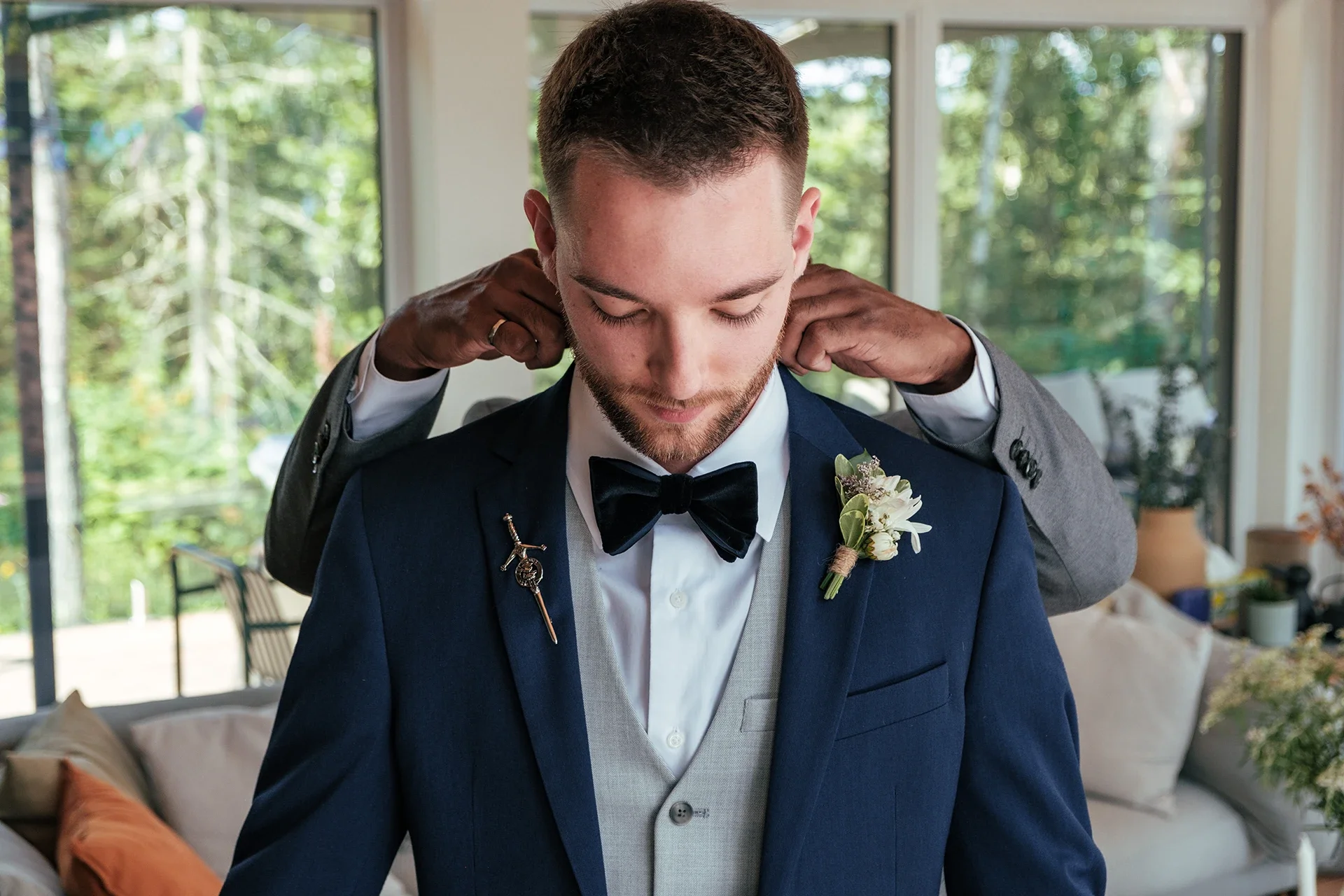 A groom in a navy blue suit and bow tie getting ready with help adjusting his attire during his wedding preparations in a bright room with large windows and green outdoor scenery.