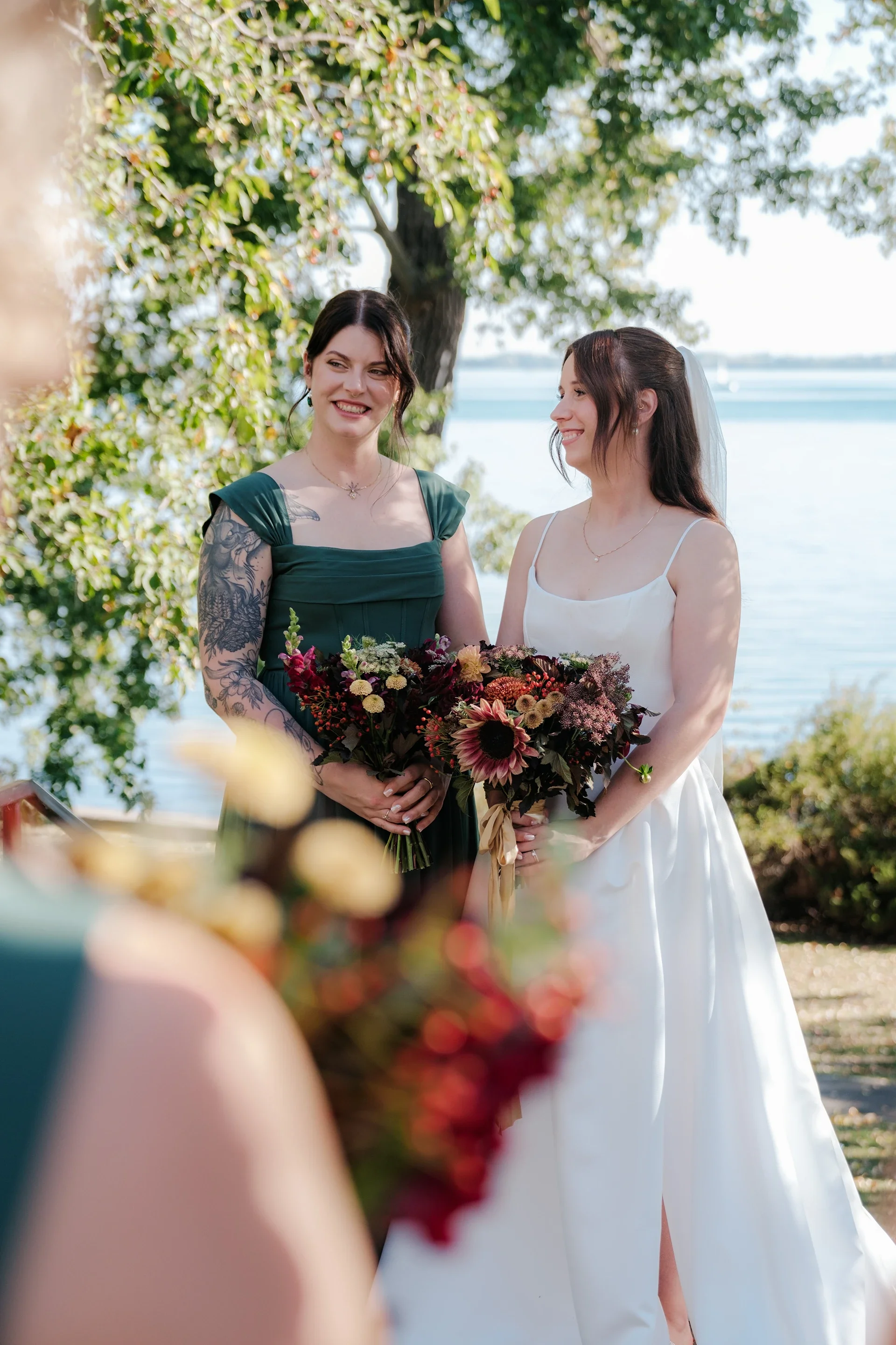 Two women, one wearing a white dress and the other in a dark green dress with tattoos on her arm, standing outdoors by the water under a tree, both holding bouquets of flowers and smiling at each other during a wedding.