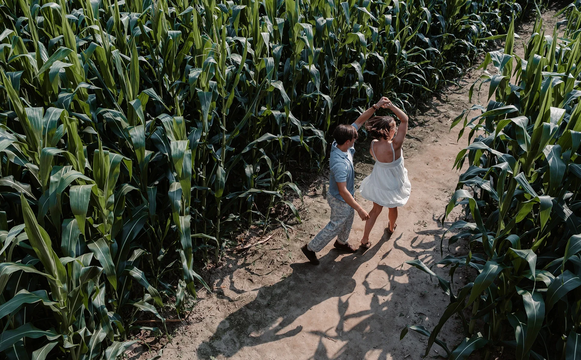 A young boy and girl dancing hand in hand on a dirt path through a cornfield, casting shadows on the ground.