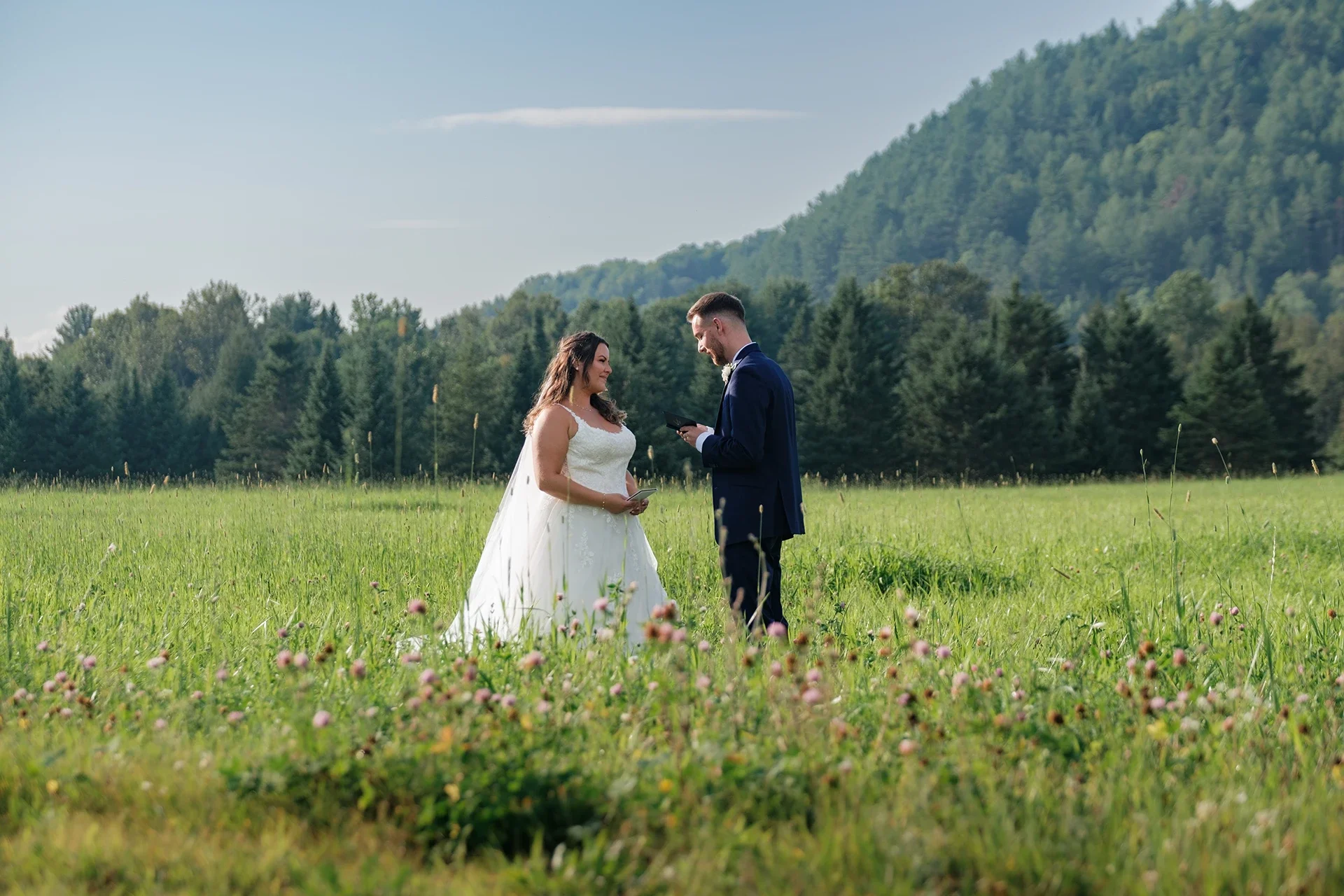 A bride and groom standing in a grassy field with mountains and trees in the background, during their wedding ceremony.