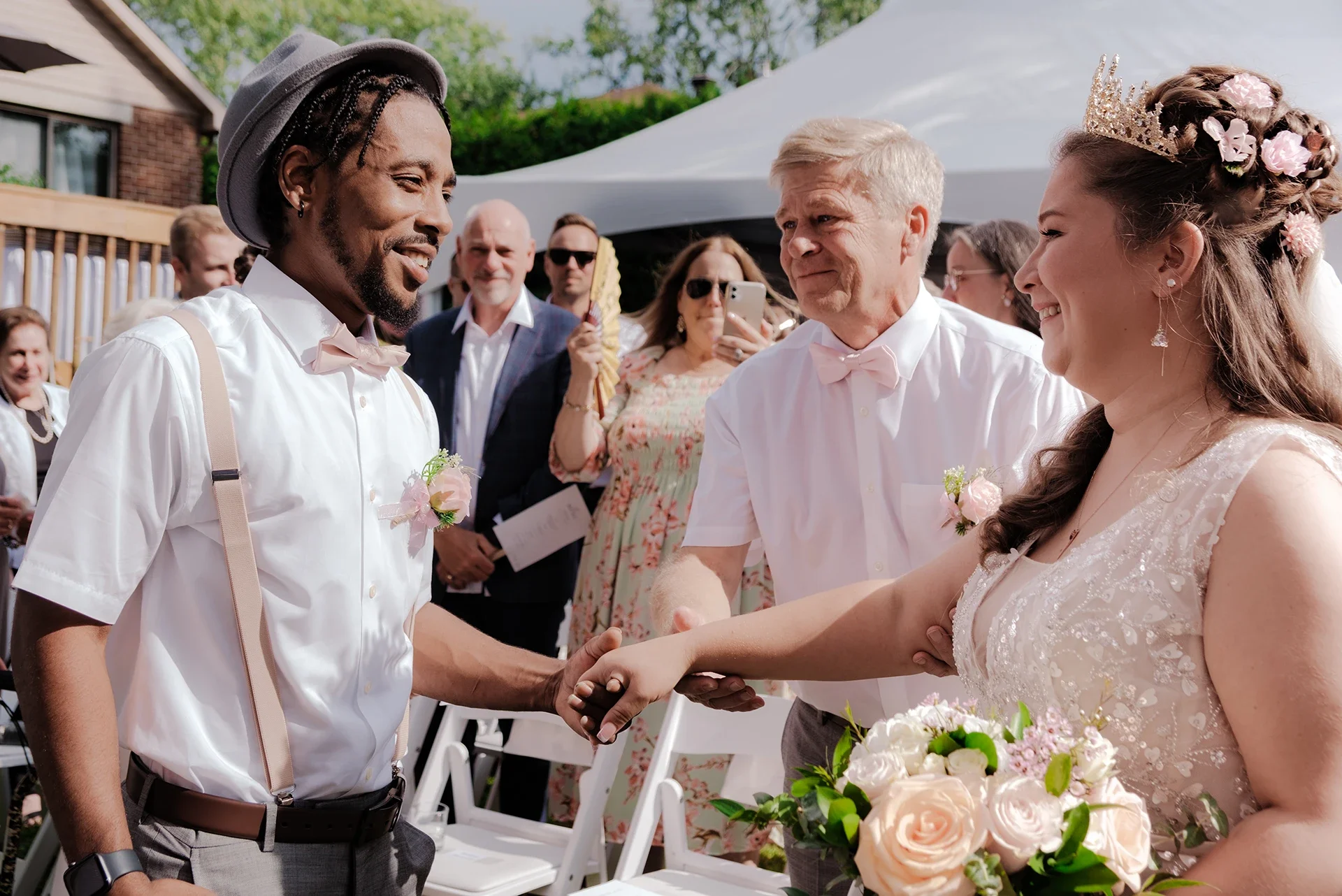 A wedding ceremony outdoors where a groom in suspenders and a bow tie is shaking hands with the bride, who is holding a bouquet and wearing a flower crown and dress. Guests in the background are capturing the moment.