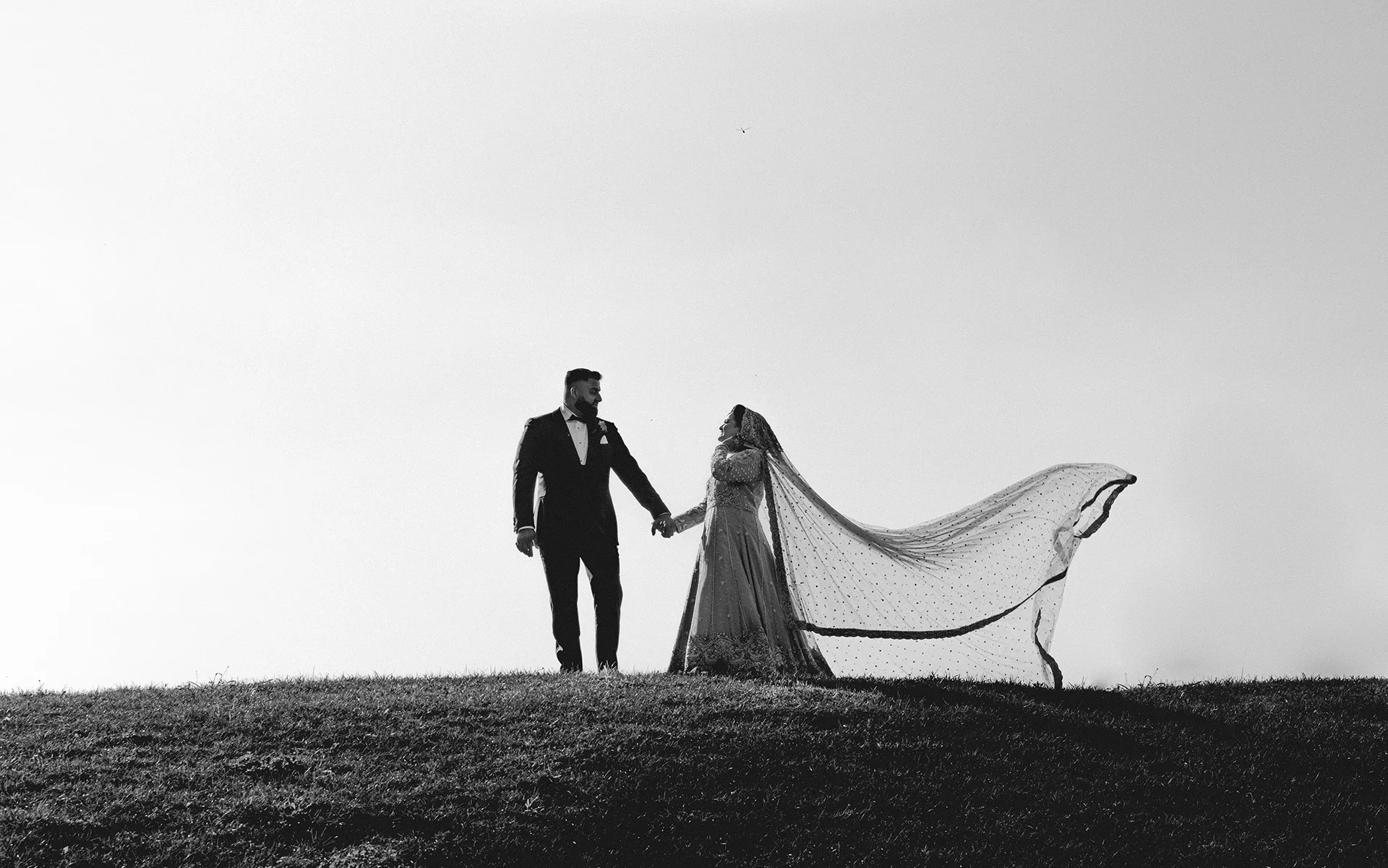 Black and white photo of a bride and groom holding hands on a grassy hill, with the bride's veil flowing in the wind