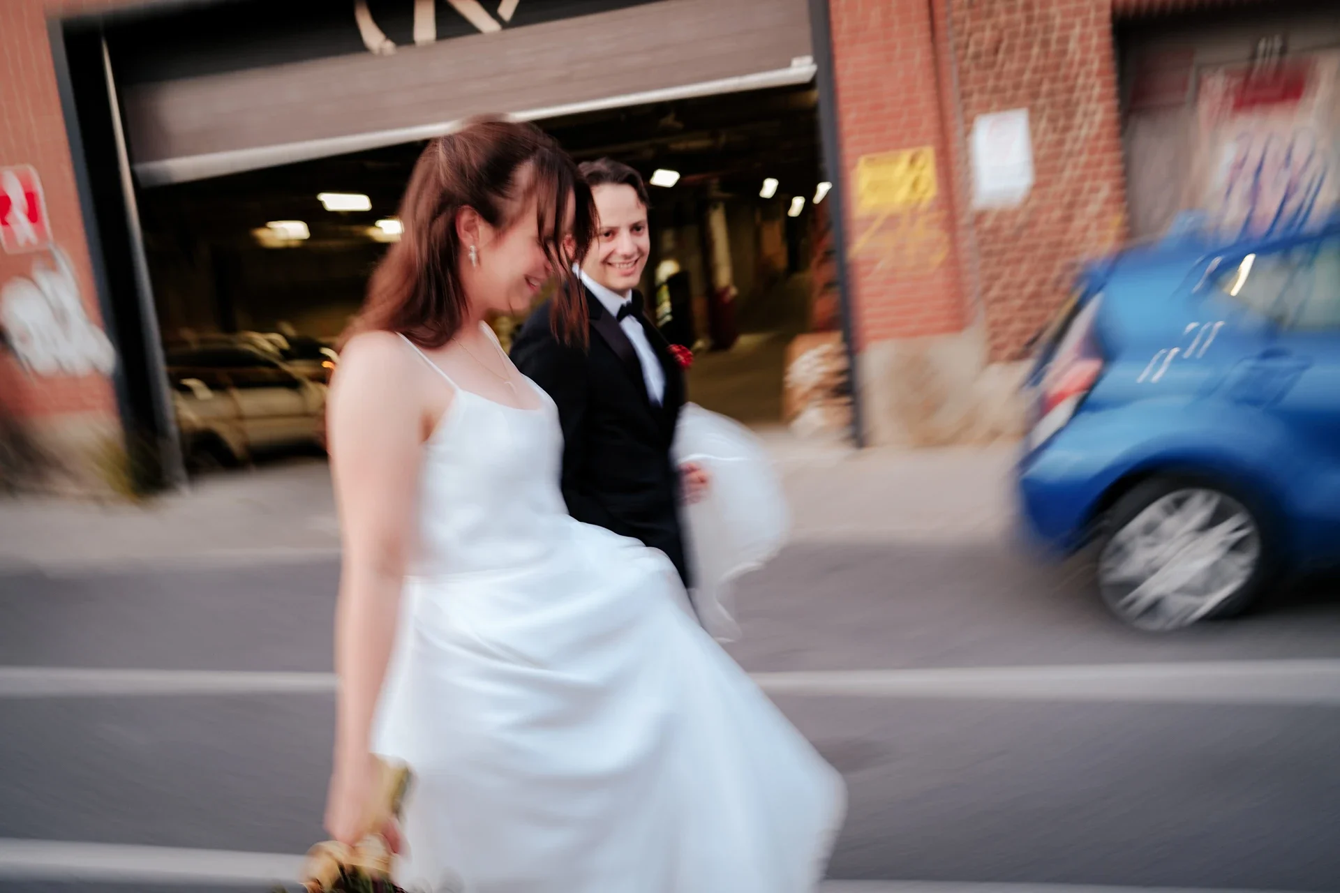 A woman in a white wedding dress and a man in a black tuxedo walking outside, smiling.