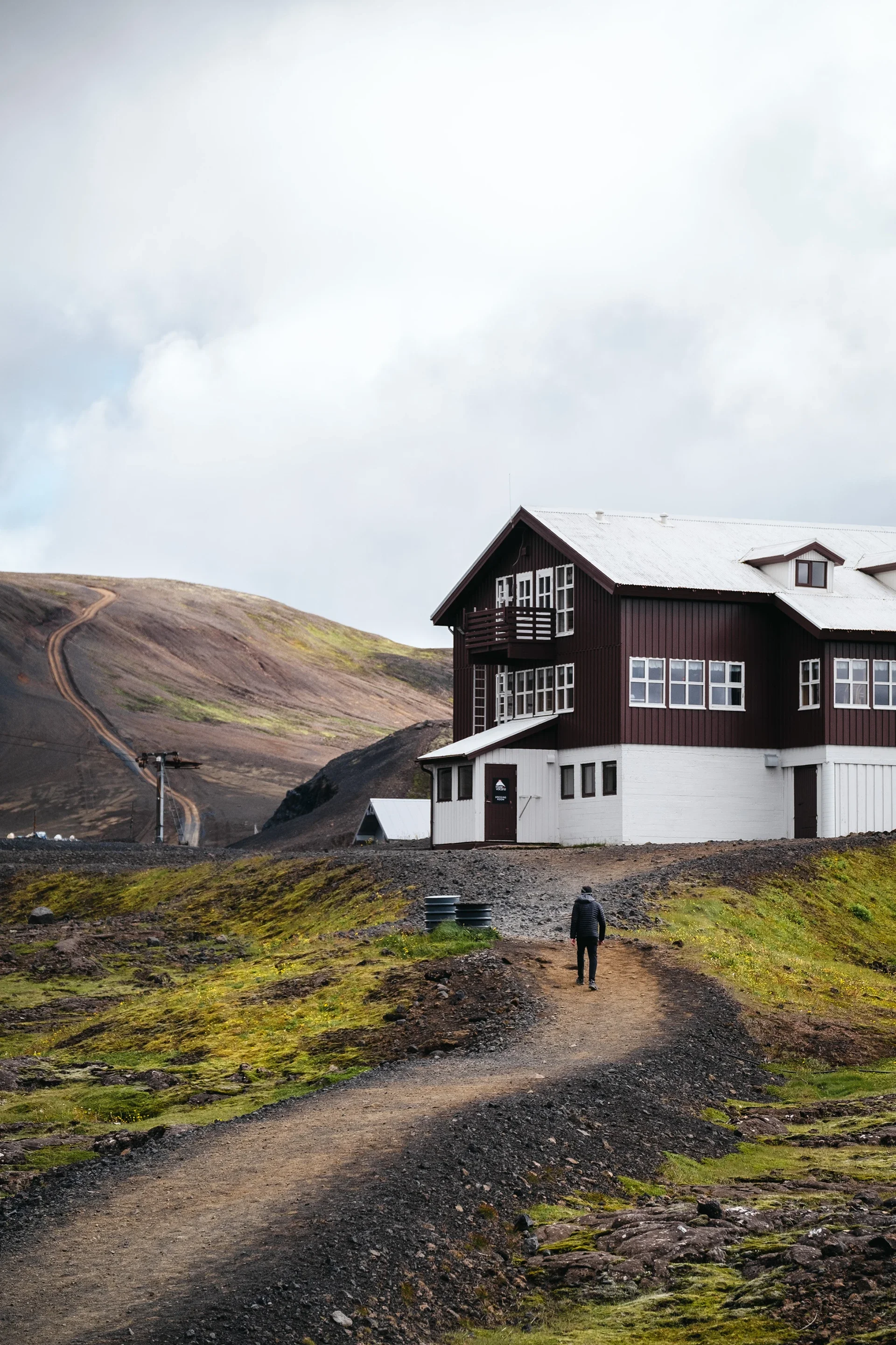 A person walking on a dirt path towards a large two-story house with dark brown and white siding, situated on a hilly landscape with green moss and rocky terrain outside.