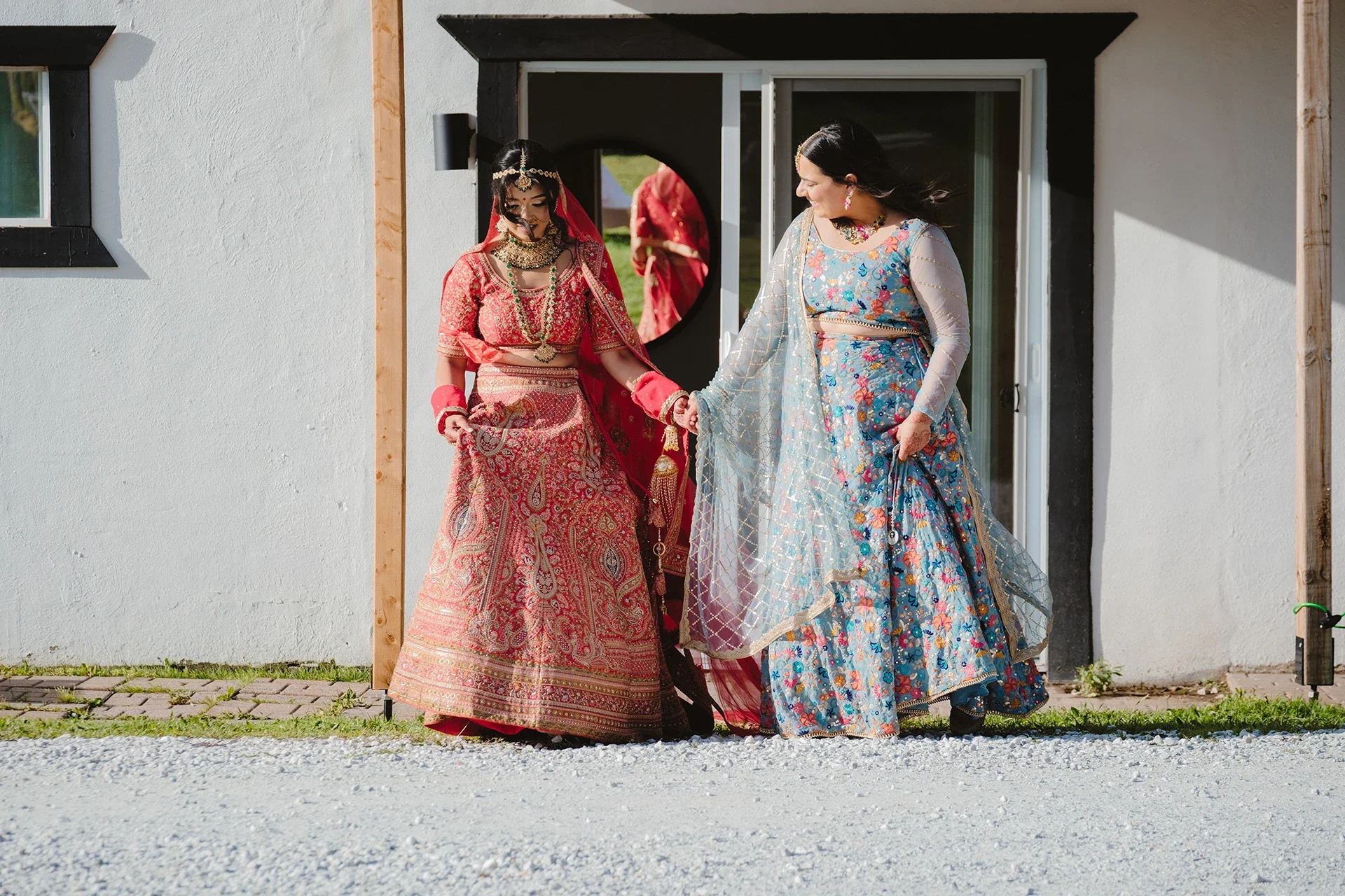 Two women in colorful traditional Indian wedding attire holding hands outside a white building.