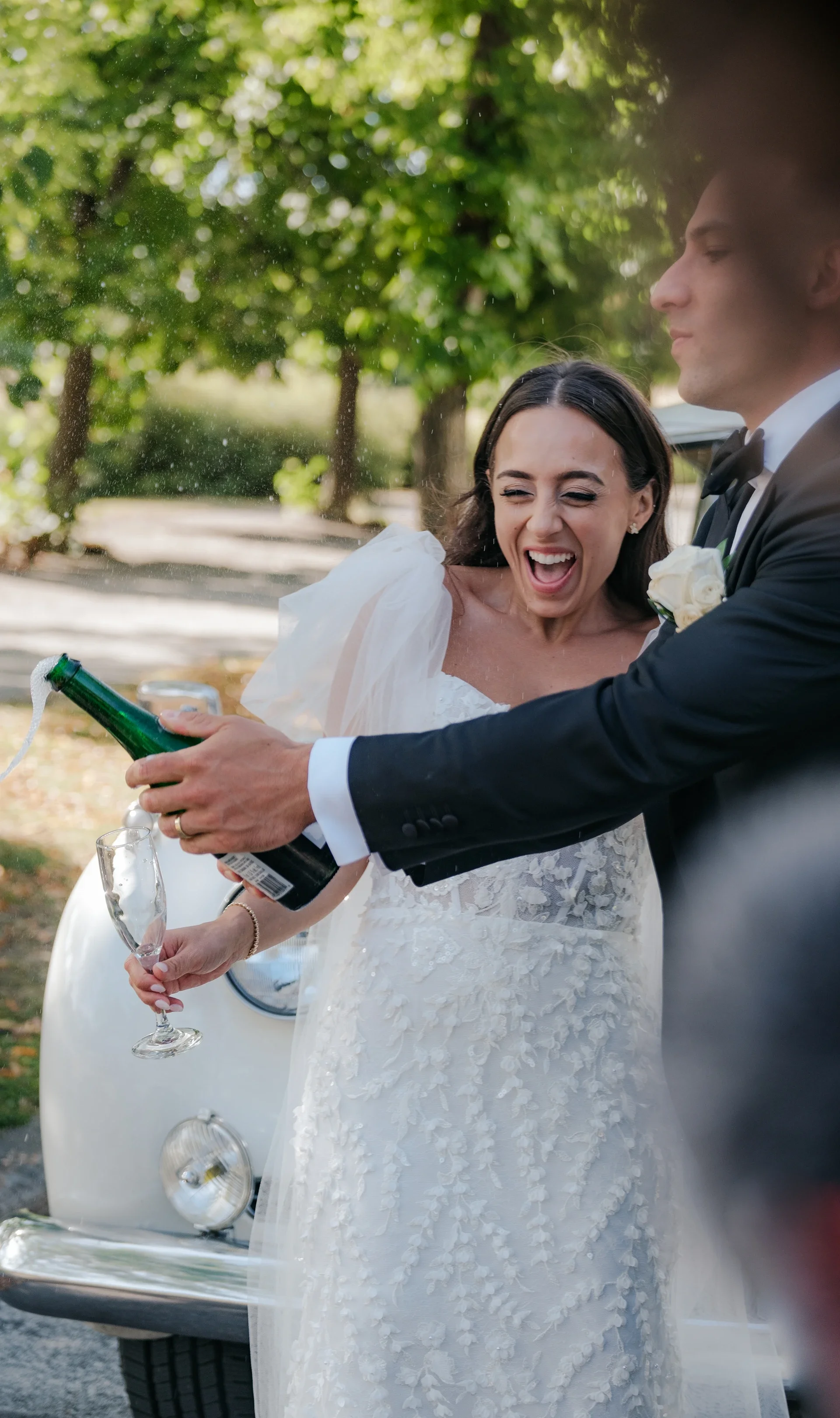 Bride and groom in wedding attire opening a bottle of champagne together outdoors with trees in the background.