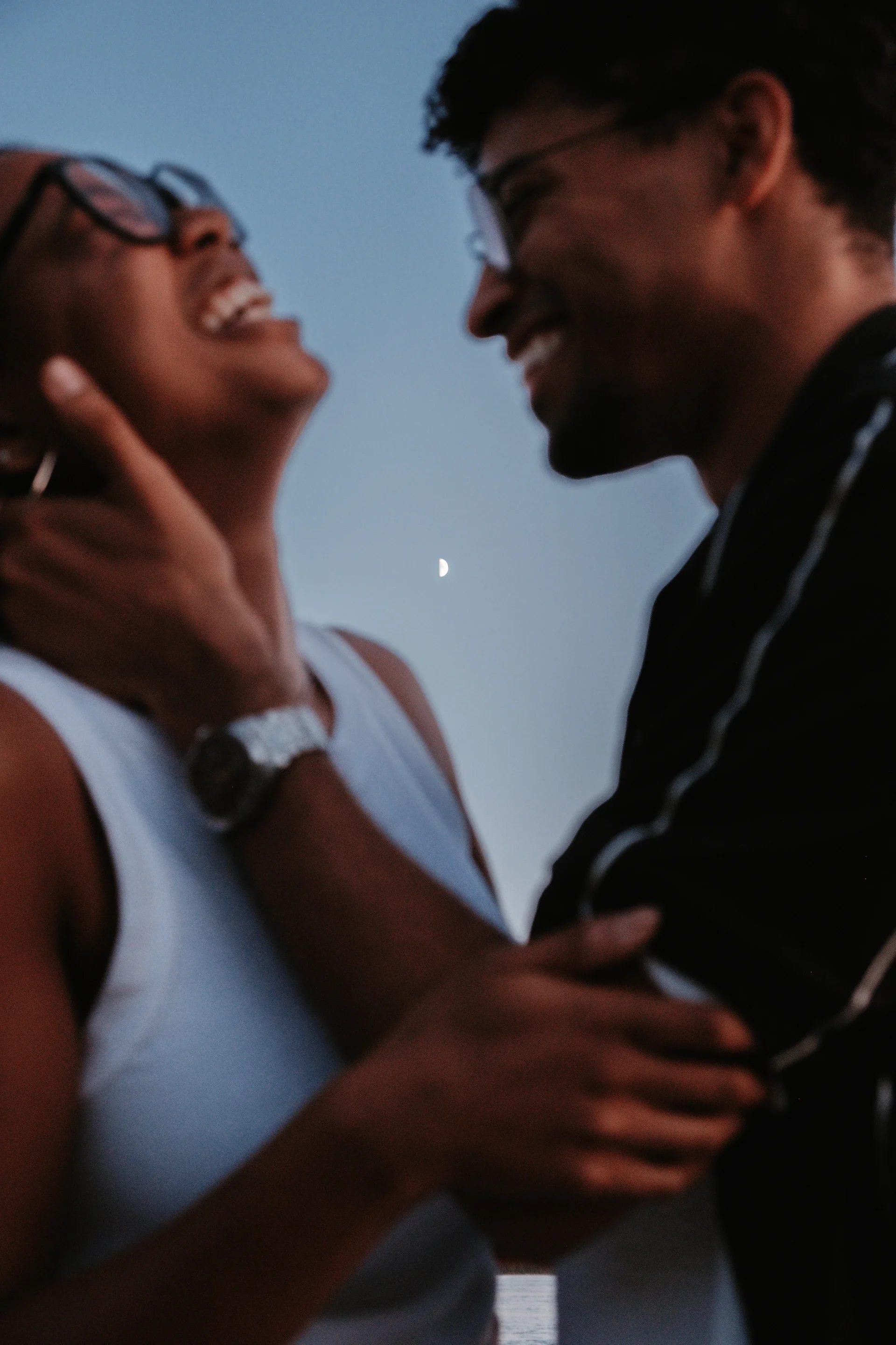 A couple laughing and smiling at each other outdoors during dusk, with the moon visible in the background sky.