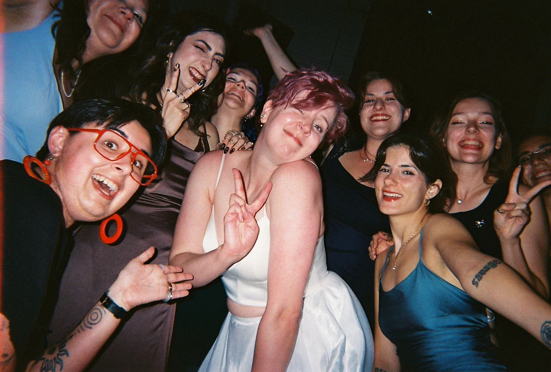 Group of women at a party or celebration smiling and posing for the camera, with some making peace signs.