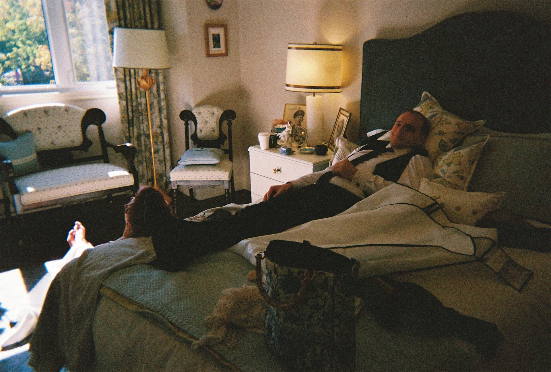 A man lying in bed and a woman sitting at the edge of the bed, holding his hand, in a bedroom with a window, chairs, a standing lamp, and a nightstand with various items.