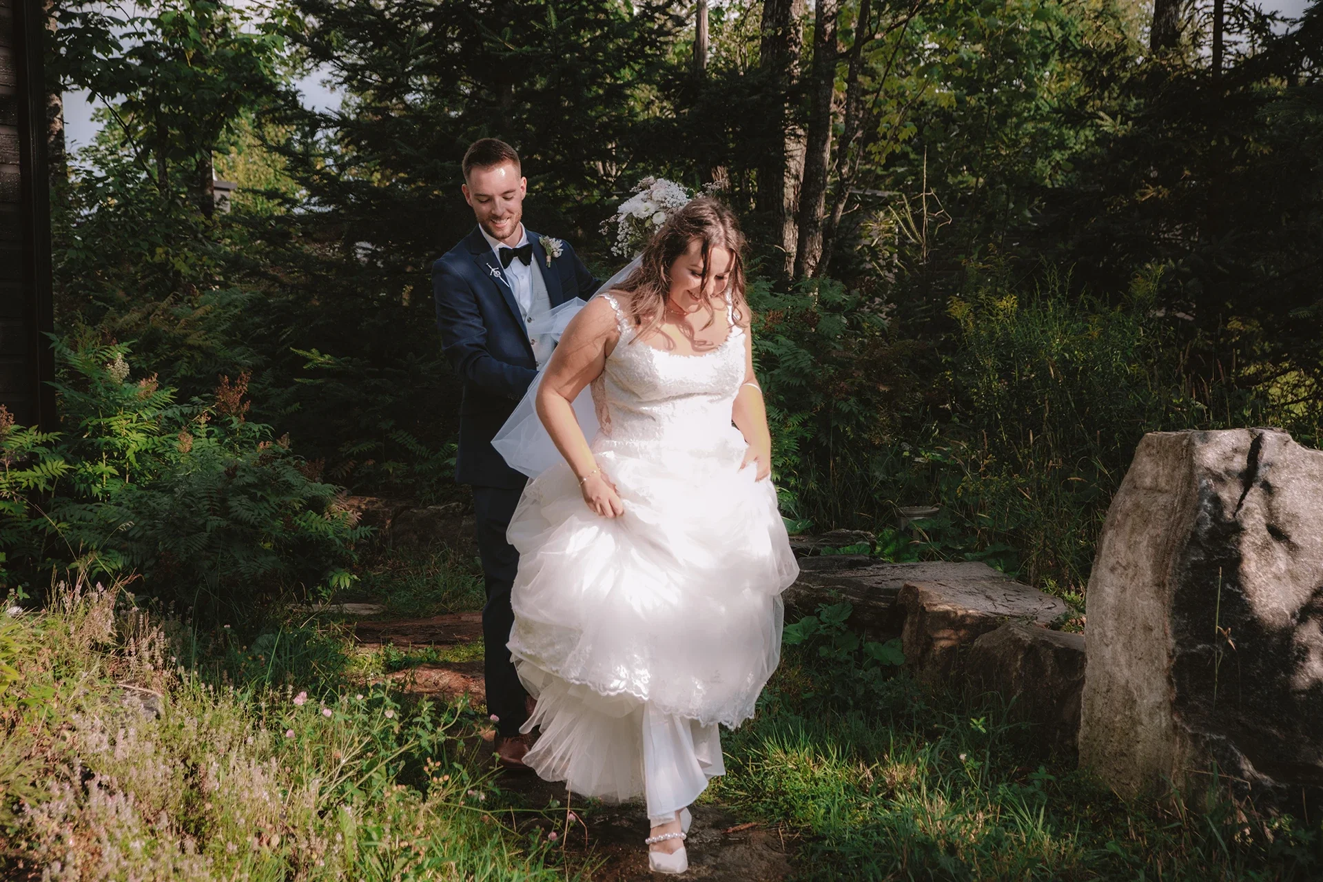 A bride in a white wedding dress and a groom in a dark suit with a bow tie outside on a wooded area, with the groom helping the bride to step over a small log.