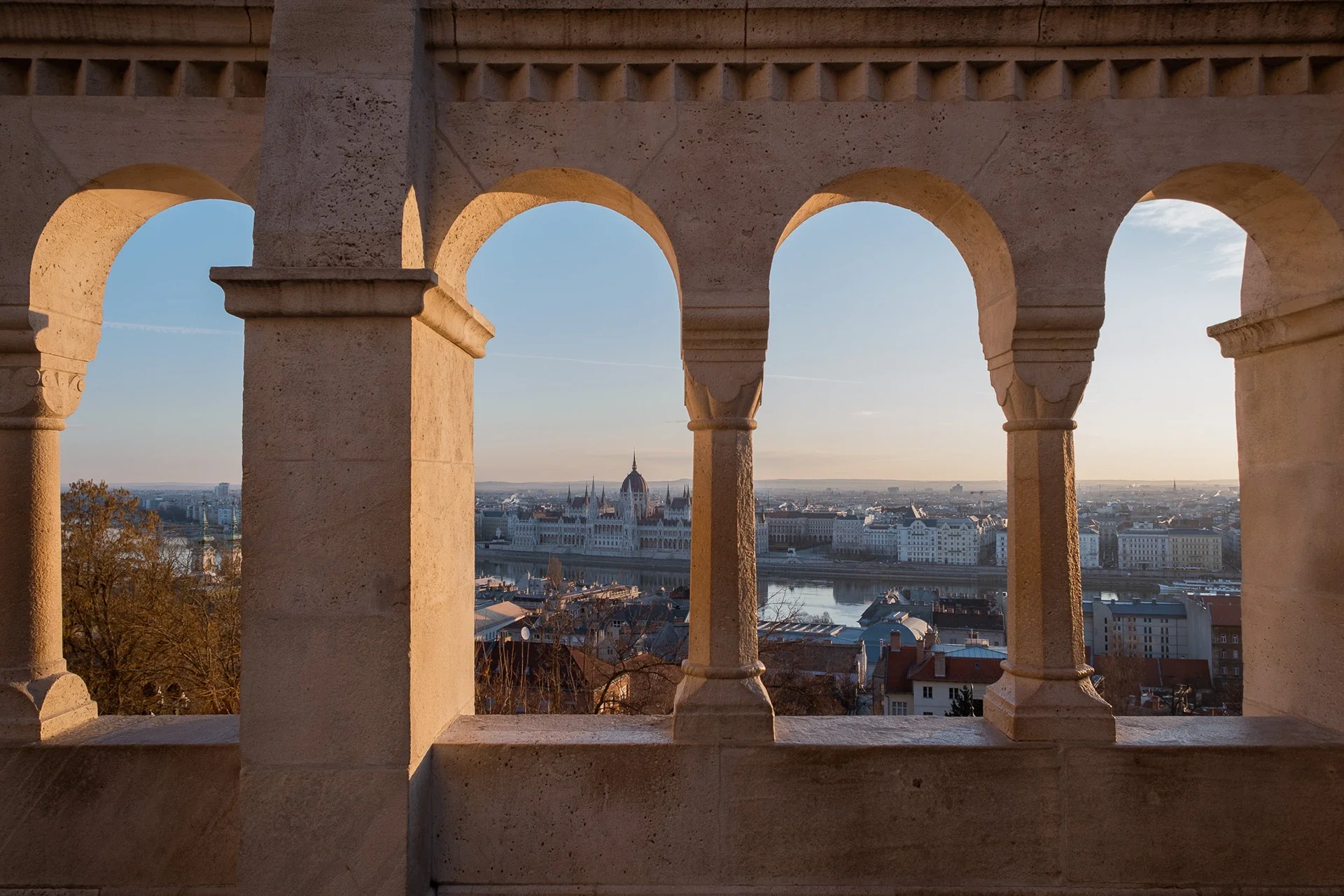 A view of Budapest, Hungary, through arched openings in a stone balcony or tower at sunset, featuring the Parliament Building along the Danube River.